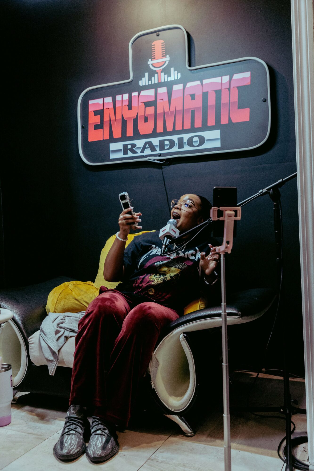 Person sitting in a chair, holding a phone, with a sign reading 'ENQUETIC RADIO' behind them.