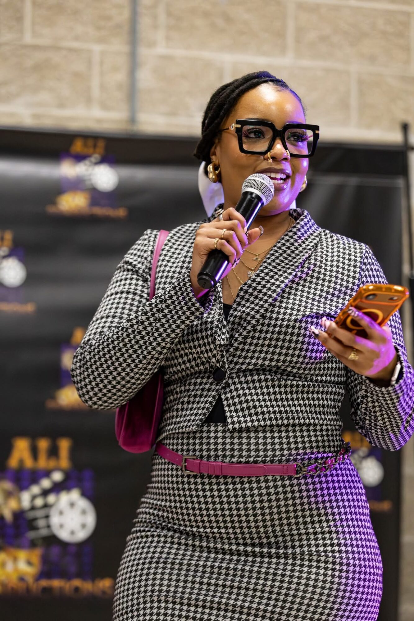 Woman with glasses holding a microphone and a phone, wearing a checkered suit, standing in front of a black backdrop.