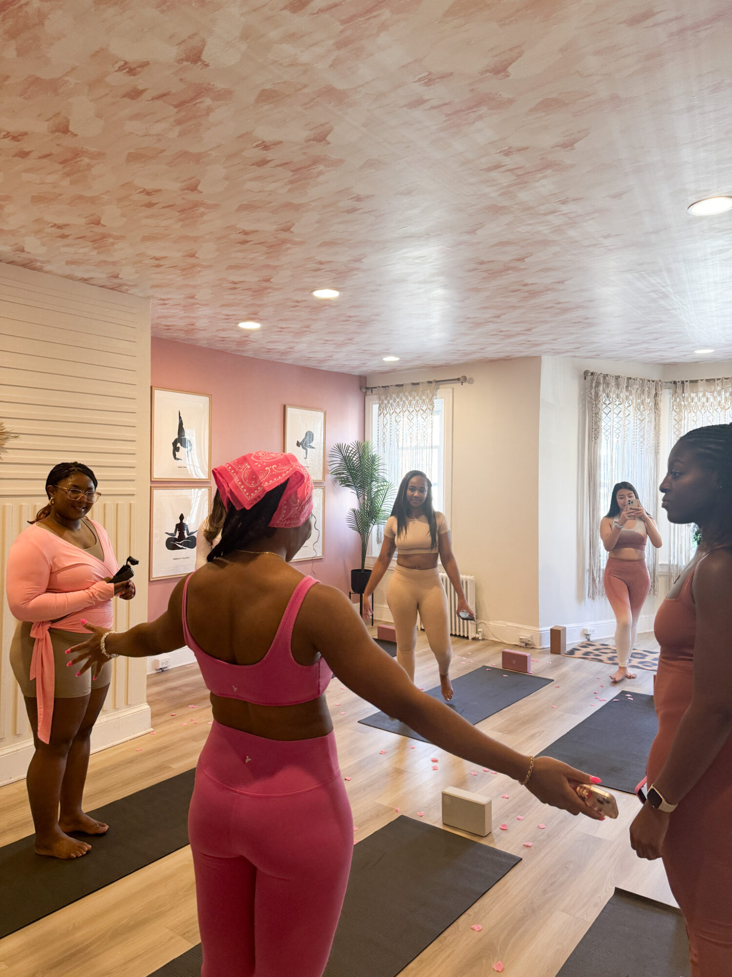 Group of women in a room with yoga mats, some holding hands, others standing, with artwork and curtains in background.