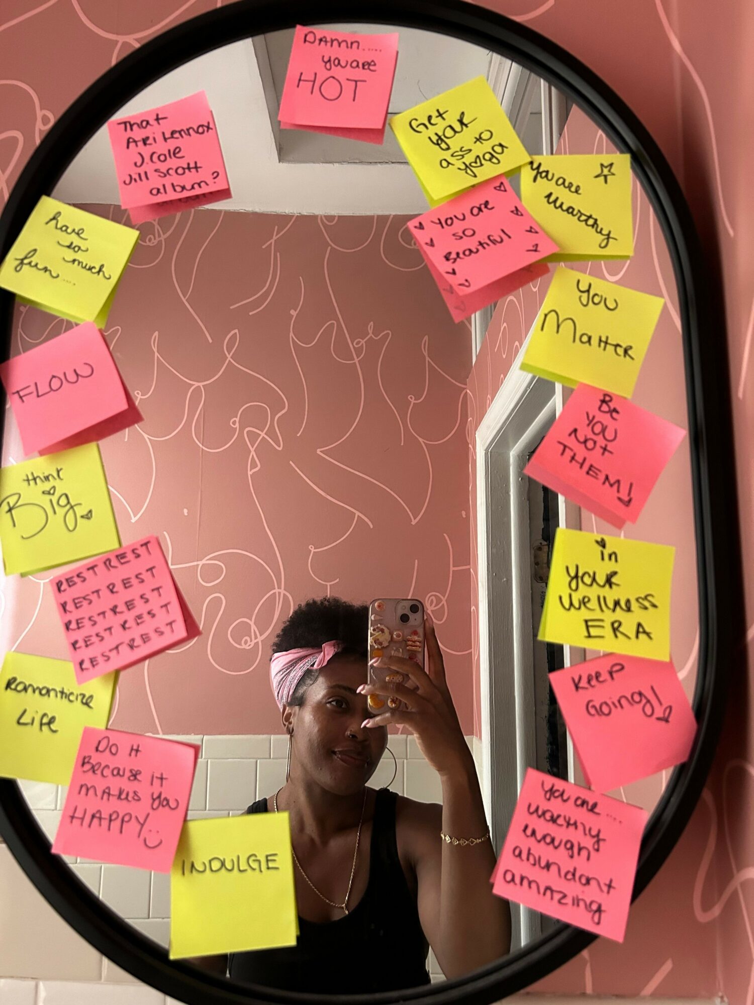 Person taking a mirror selfie with sticky notes on the mirror, pink and yellow notes with handwritten messages, pink wall background.