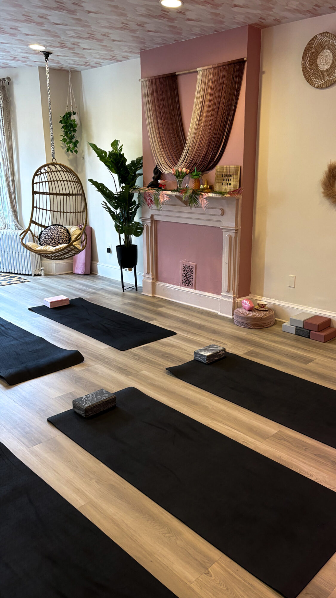 Room with yoga mats on wooden floor, wicker chair, potted plant, and decorative cabinet with curtains, in soft lighting.
