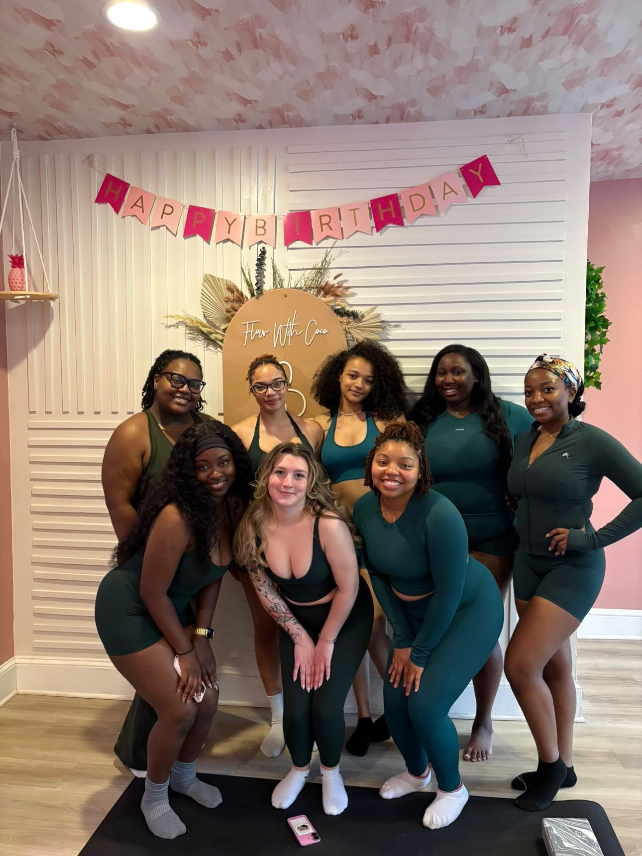 Group of eight women posing indoors with birthday decorations, smiling, some with hands on hips, in front of a white wall with pink banner.