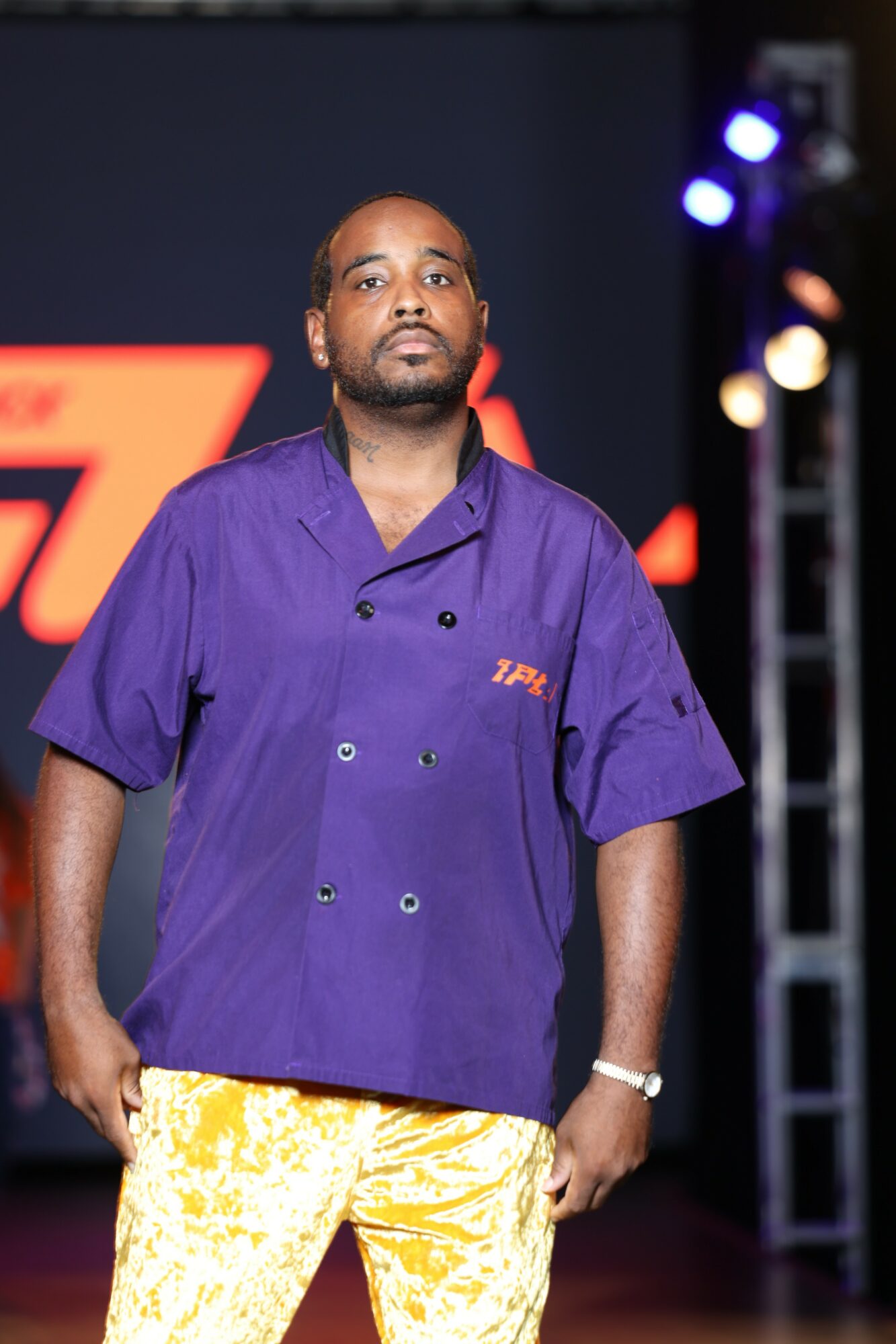 Man in purple shirt and yellow pants standing on stage with lights and a backdrop behind him.