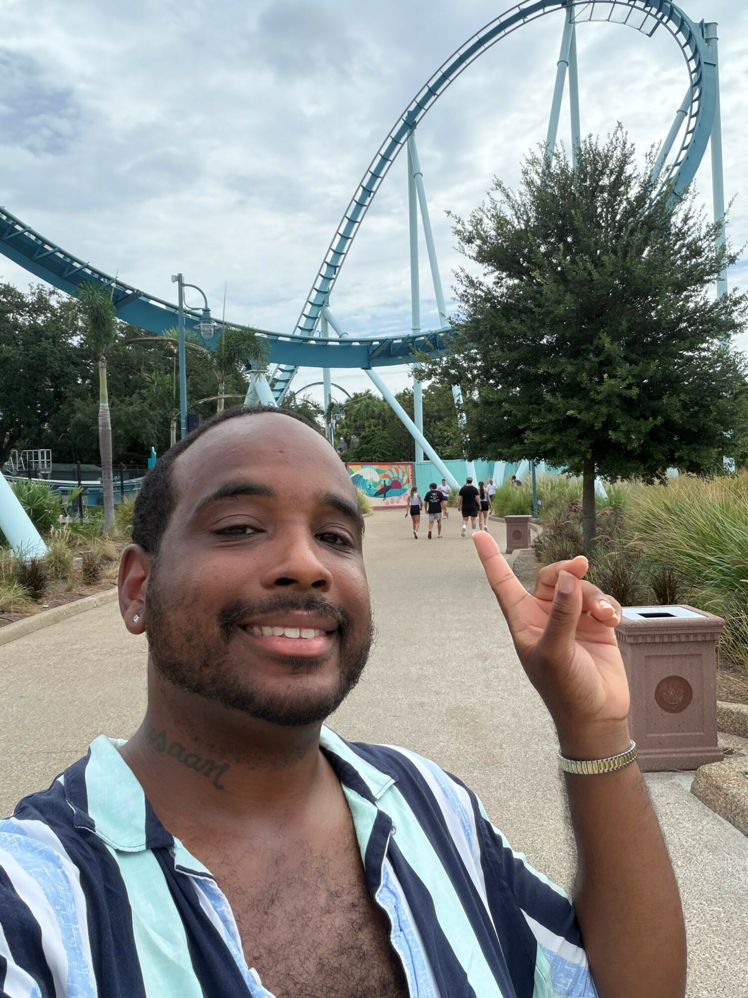 Smiling person with striped shirt pointing at a roller coaster in an amusement park, trees and a pathway in the background.