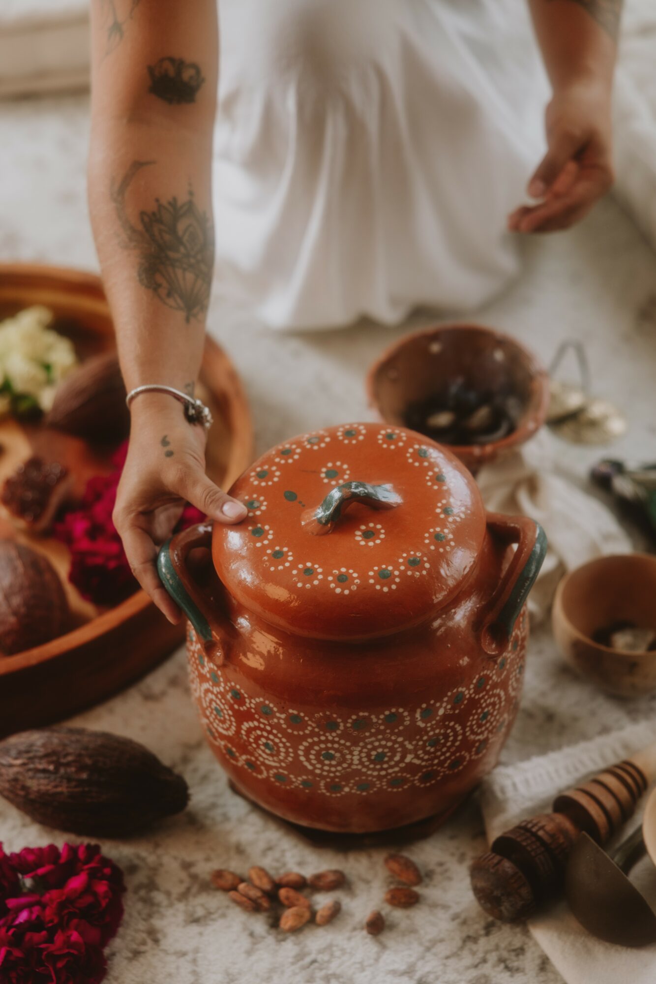 Person's hand reaching into a decorated ceramic jar surrounded by bowls and ingredients, tattoos visible on arm.