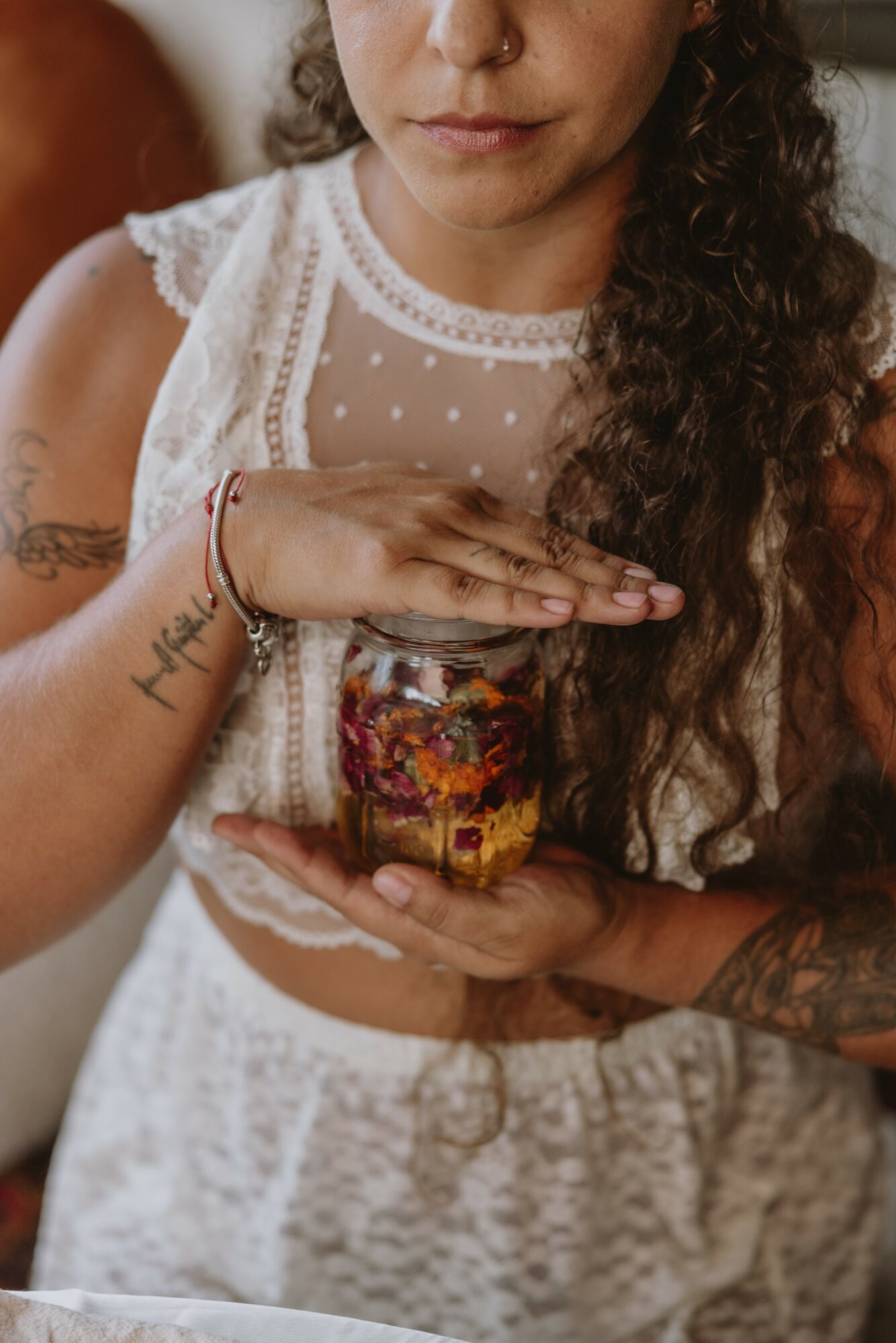 Person holding a jar filled with colorful items, wearing a white lace top and beige pants, with long curly hair.