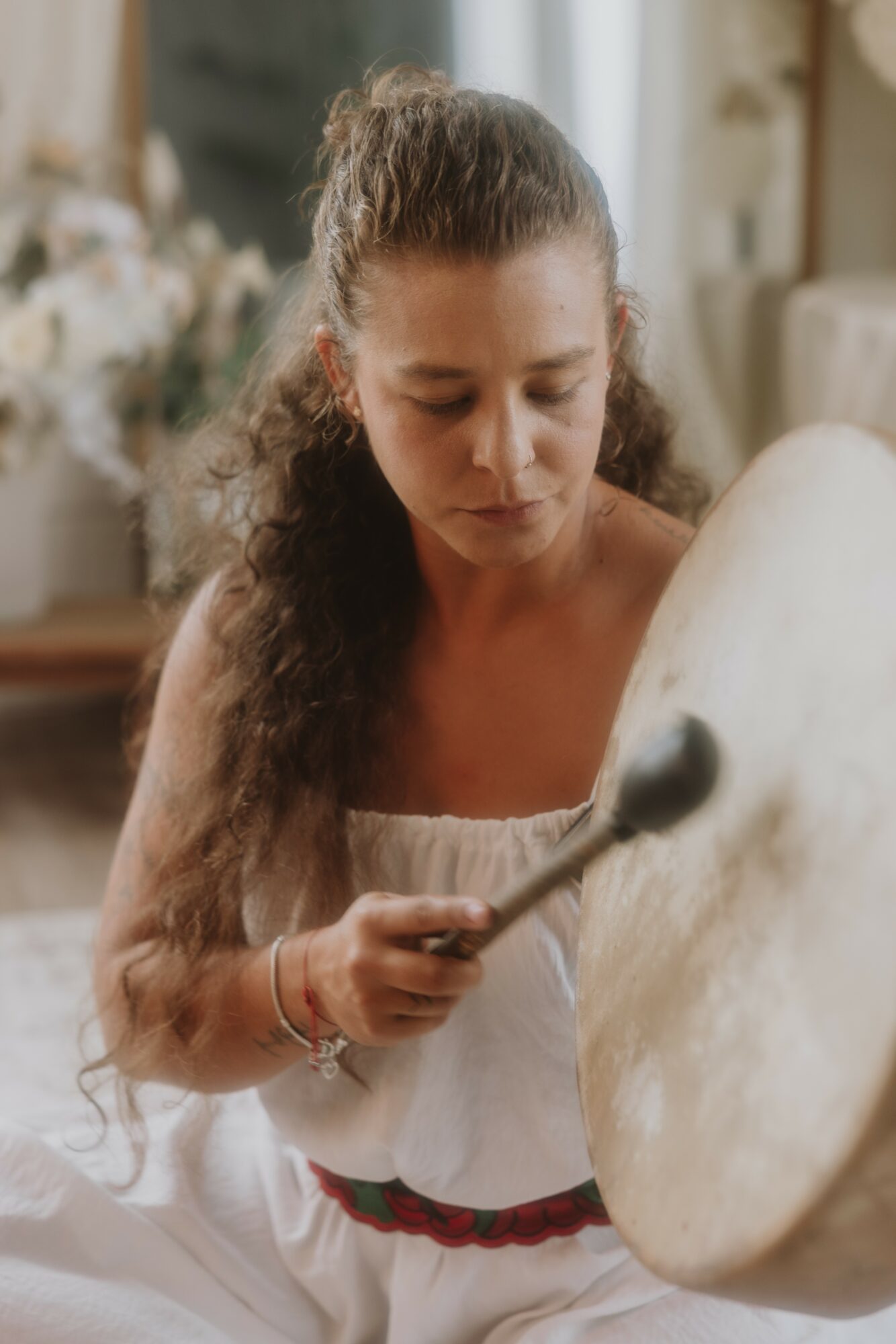 Young woman with long curly hair playing a large drum with a drumstick in a bright room.