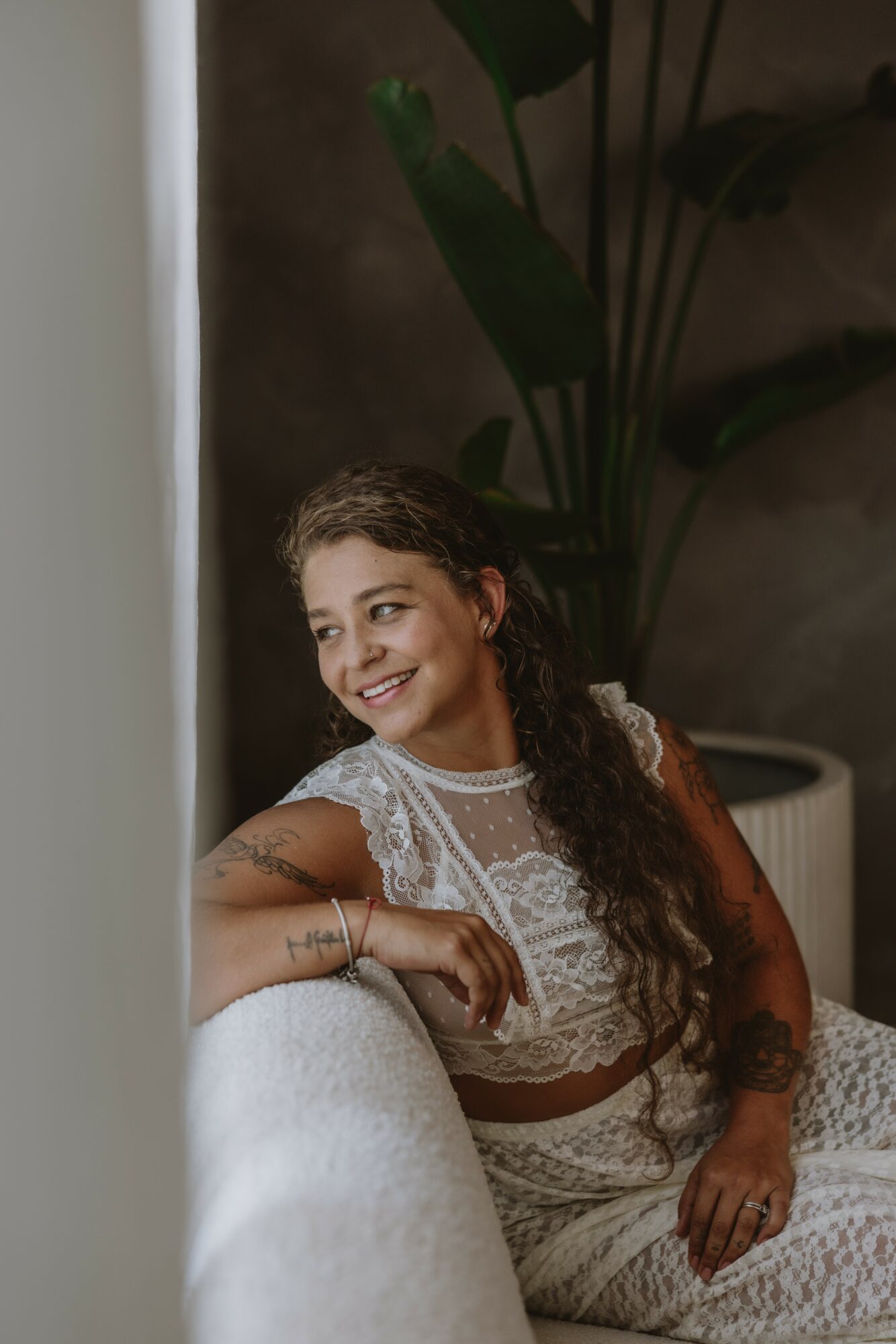 Young woman with curly hair smiling, sitting on a white couch, wearing a lace top, in a room with large plant.