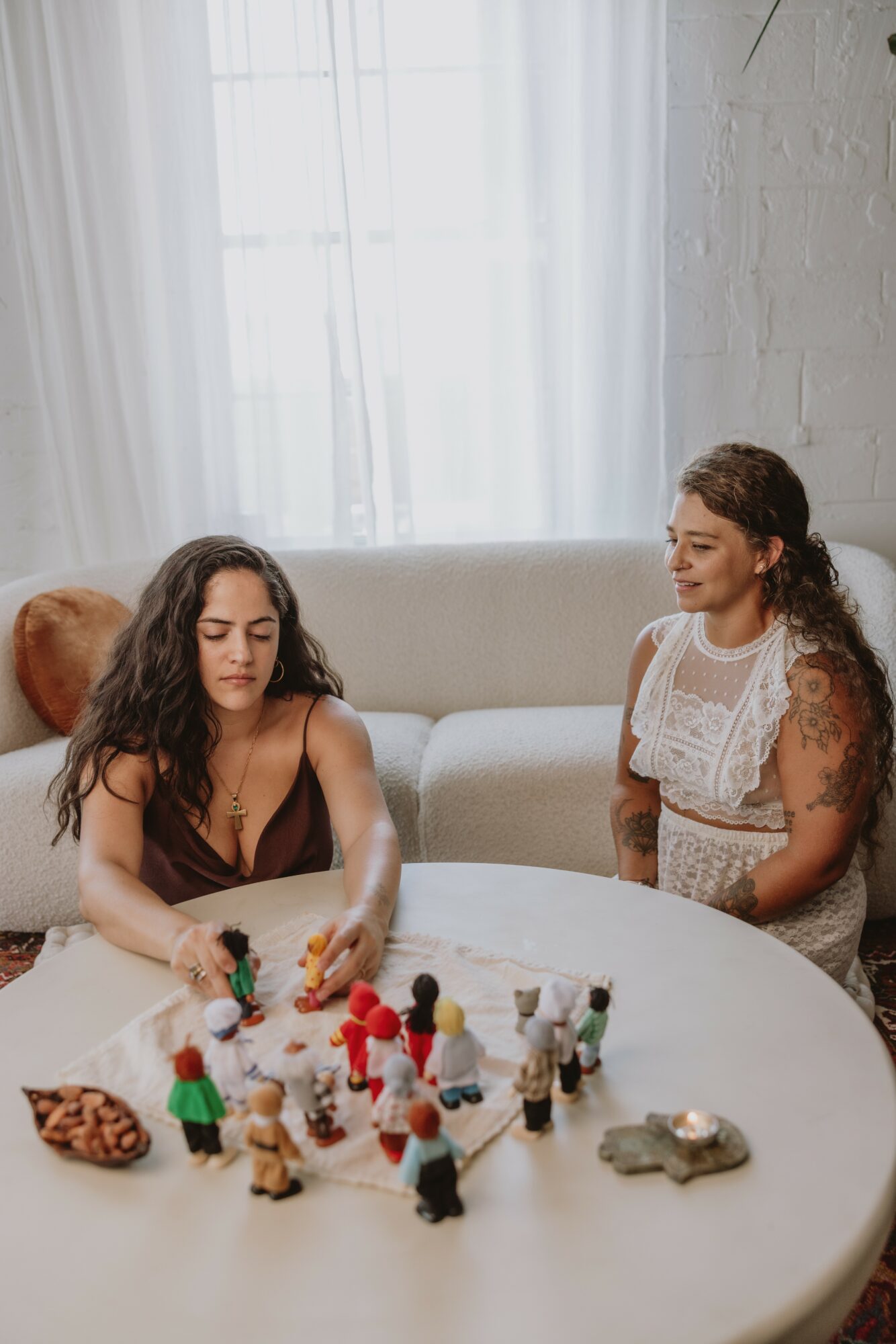 Two women playing with small figurines on a round table in a bright room with a large window.