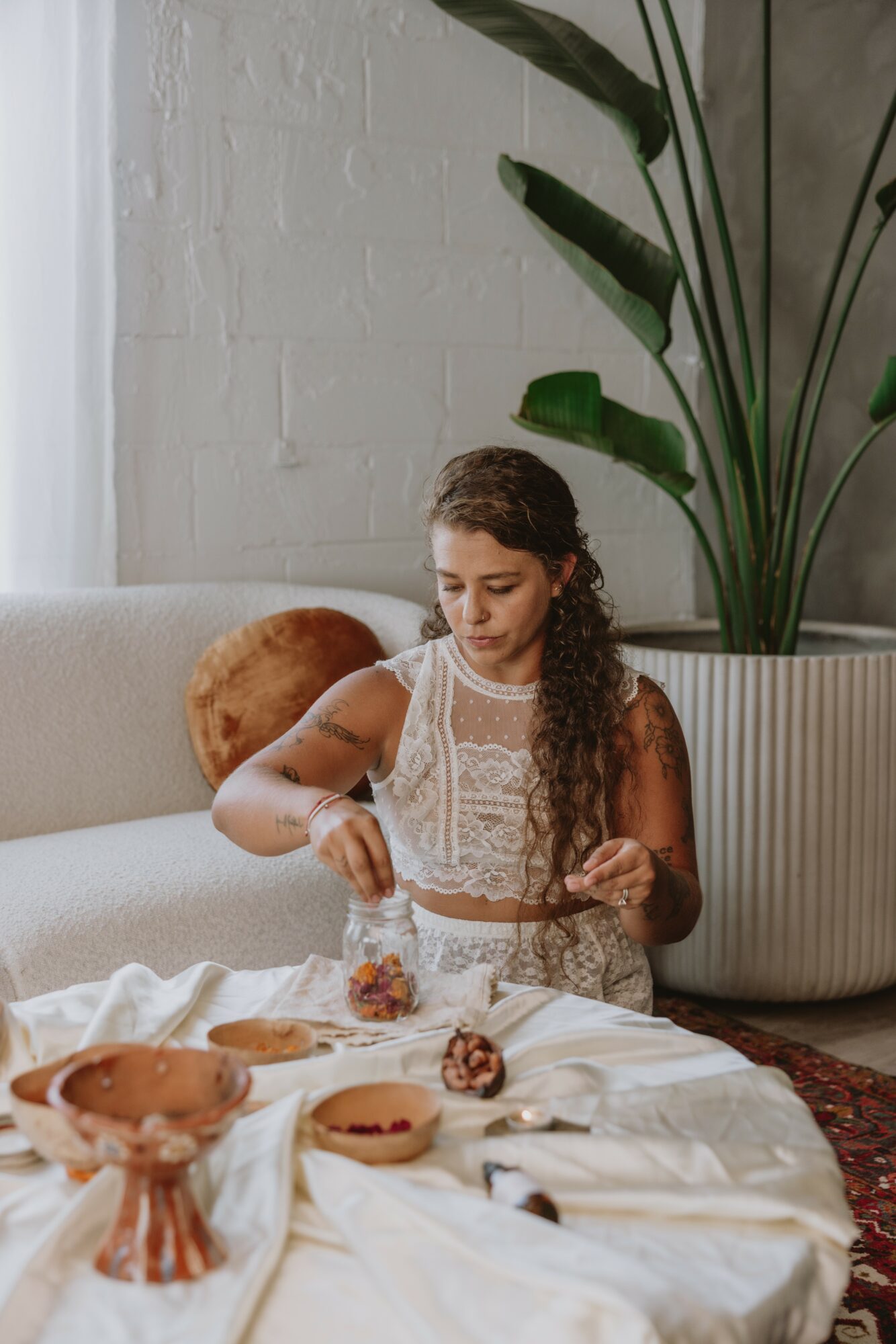 Young girl with long curly hair sitting on a sofa, preparing food on a table with cookies and a bowl, in a room with a large plant and white brick wall.