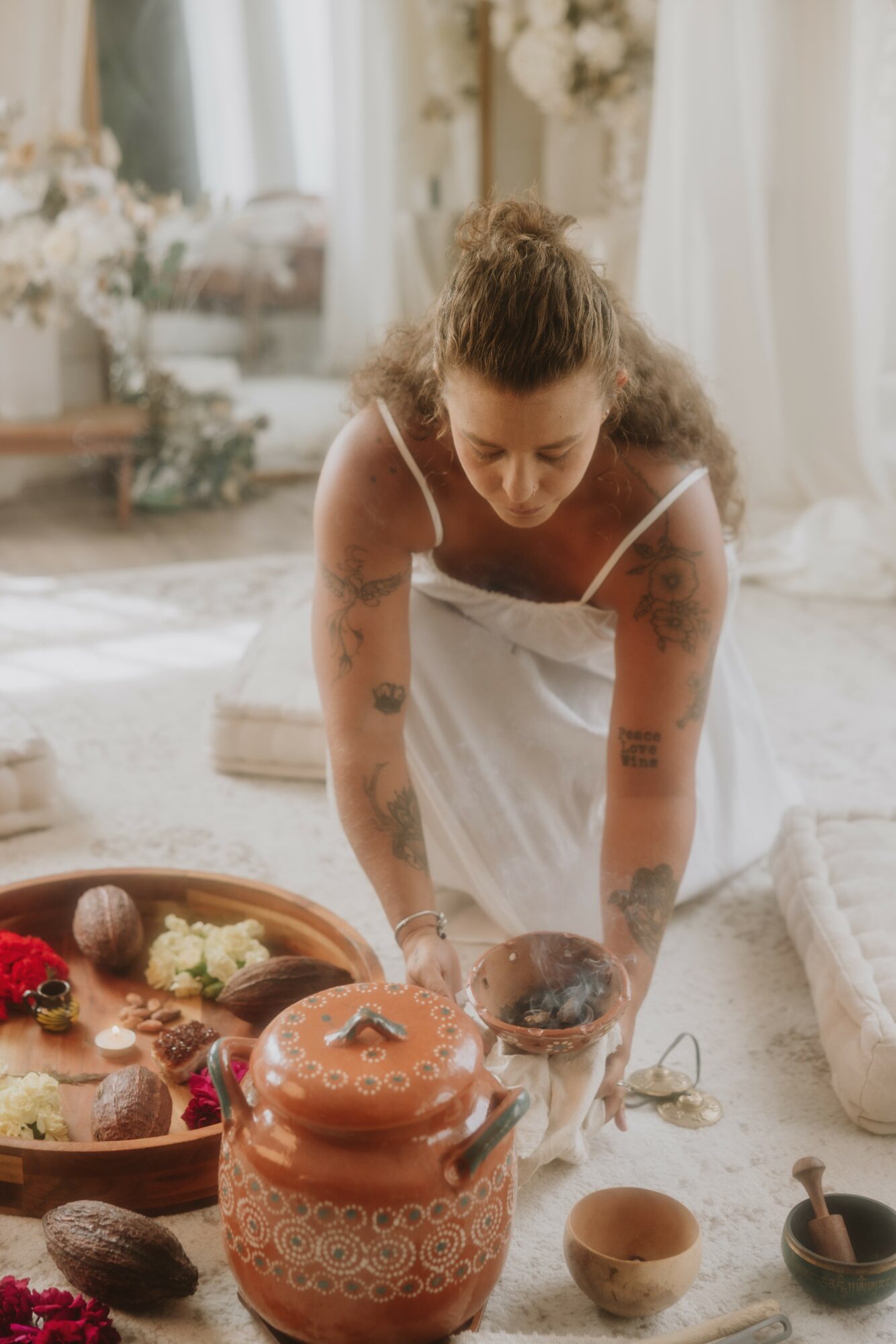 Woman with curly hair leaning forward in a bright, decorated room, surrounded by pottery and flowers.