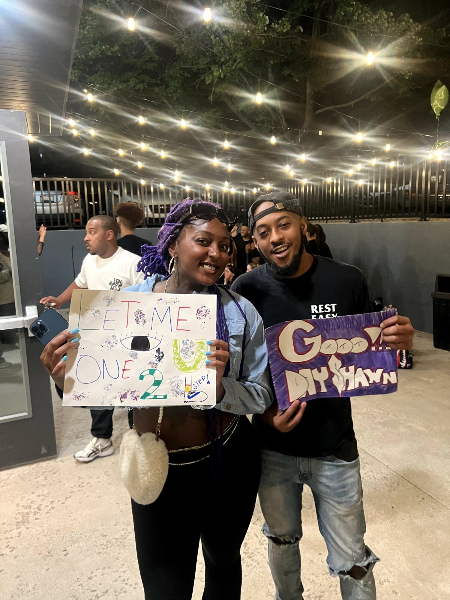 Two smiling people holding colorful signs at night, with string lights overhead and a fence in the background.