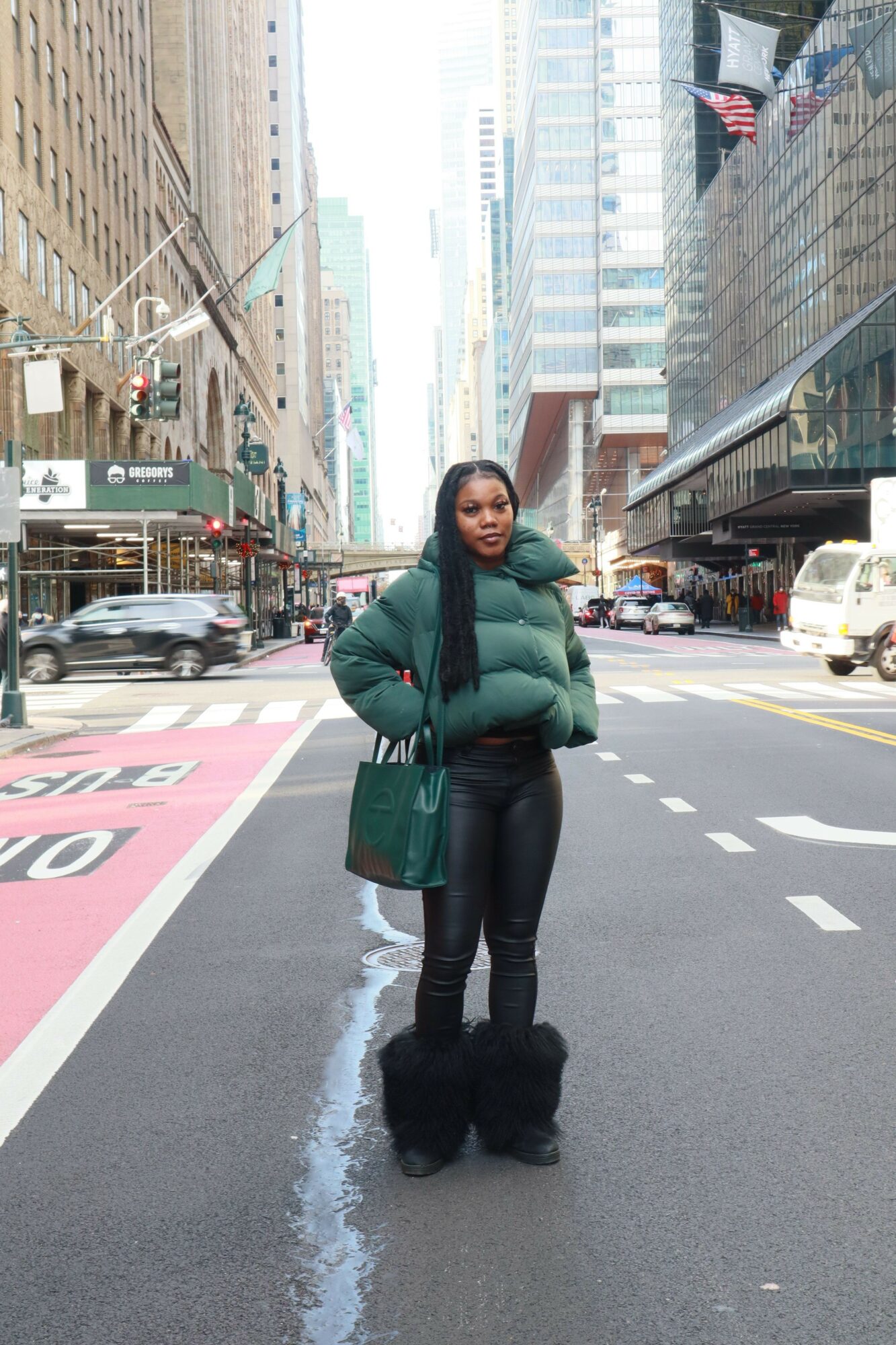 Woman standing on city street with buildings and vehicles in background, wearing green jacket and black pants.