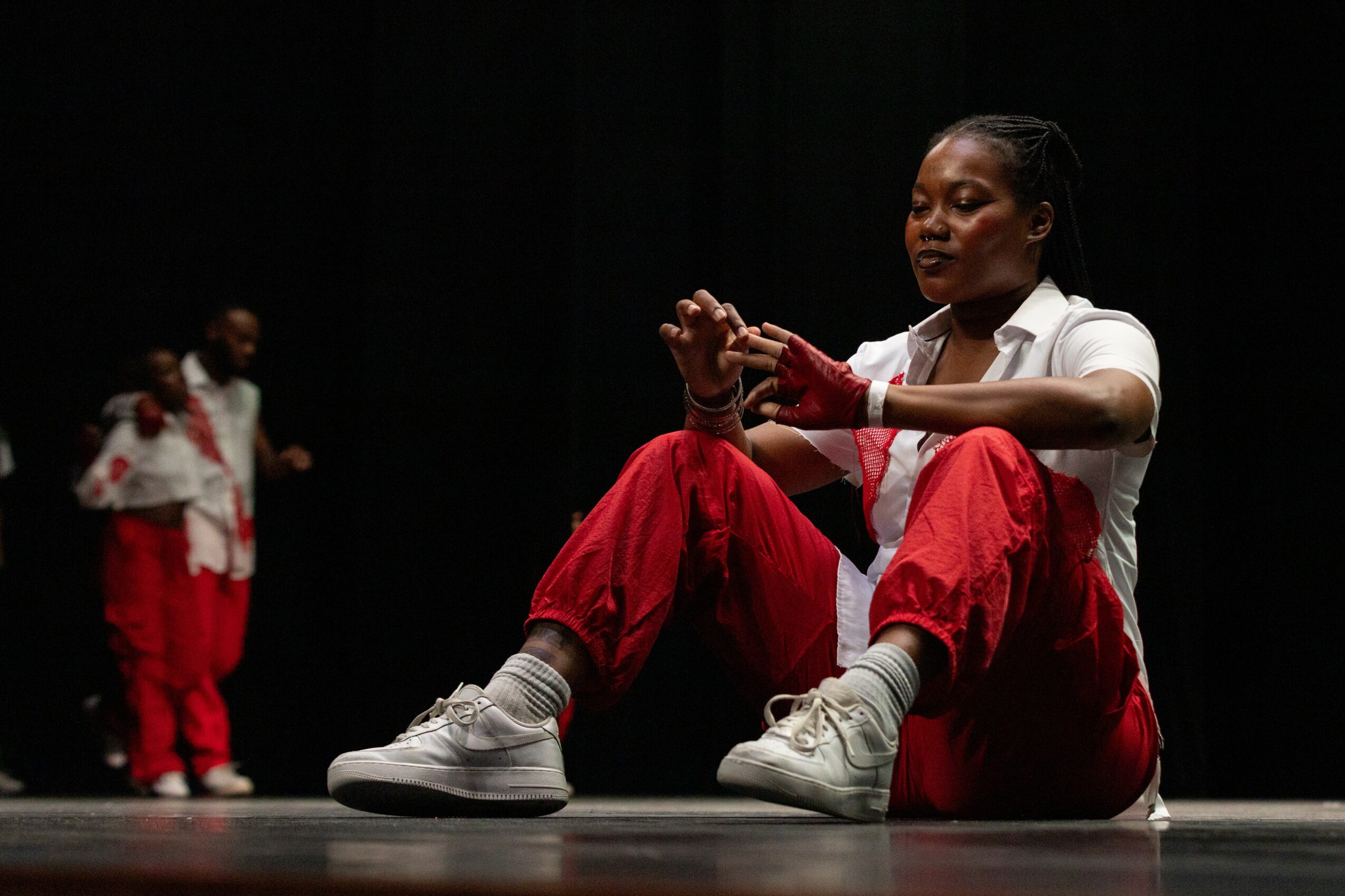 A young girl sits on the stage with her legs crossed, holding her hands together, with two other children in the background.