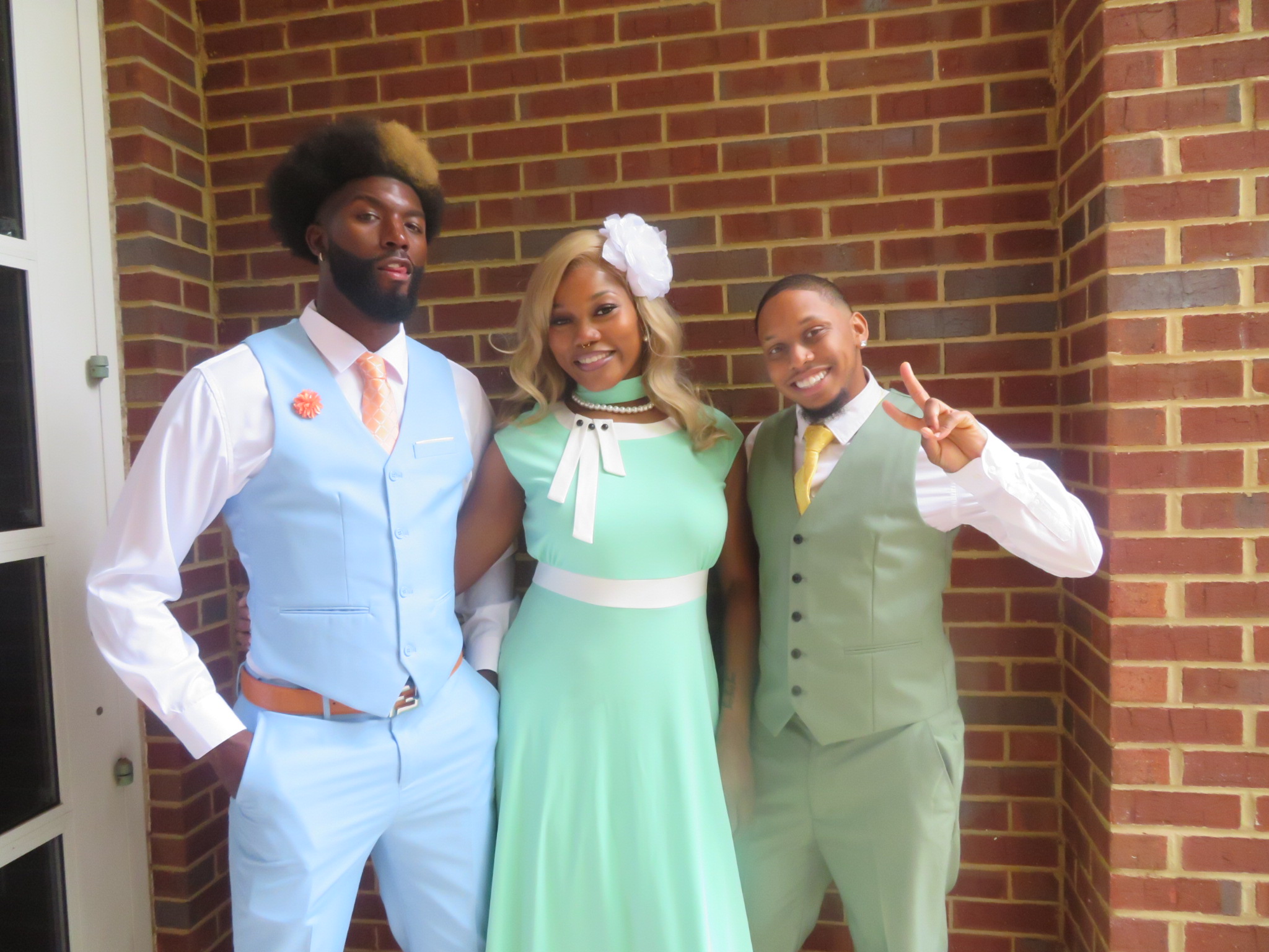 Three people standing in front of a brick wall, smiling, dressed in pastel-colored formal attire.