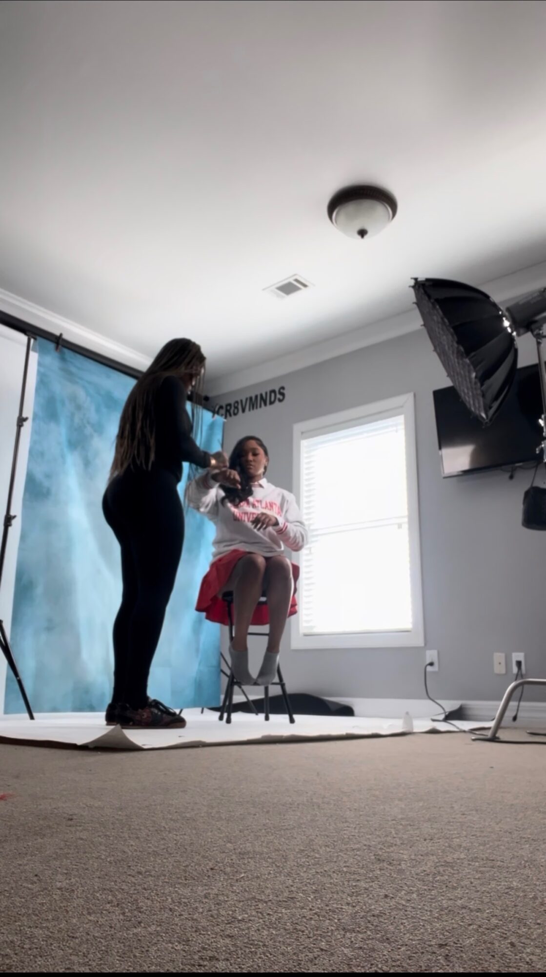 Person sitting in a chair with a makeup artist applying makeup, in a room with a window and photography equipment