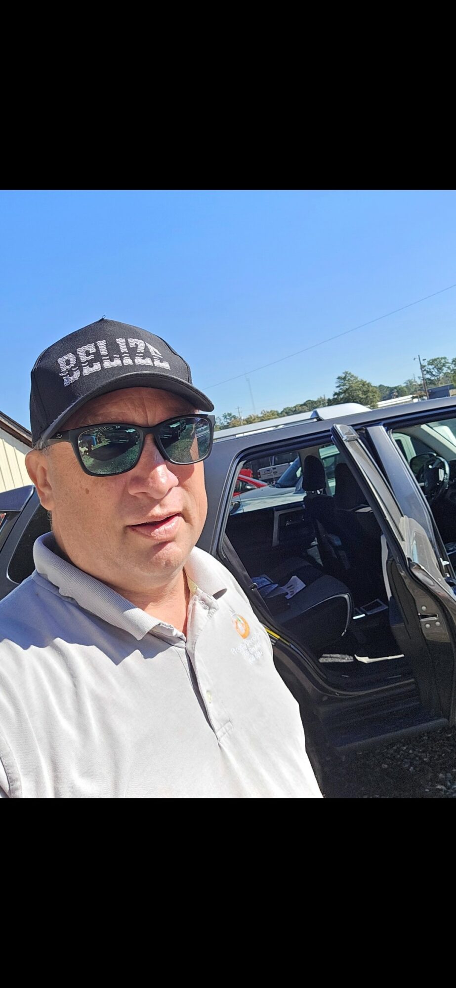 Man wearing sunglasses and a cap, standing outdoors next to a vehicle, with a clear blue sky in the background.