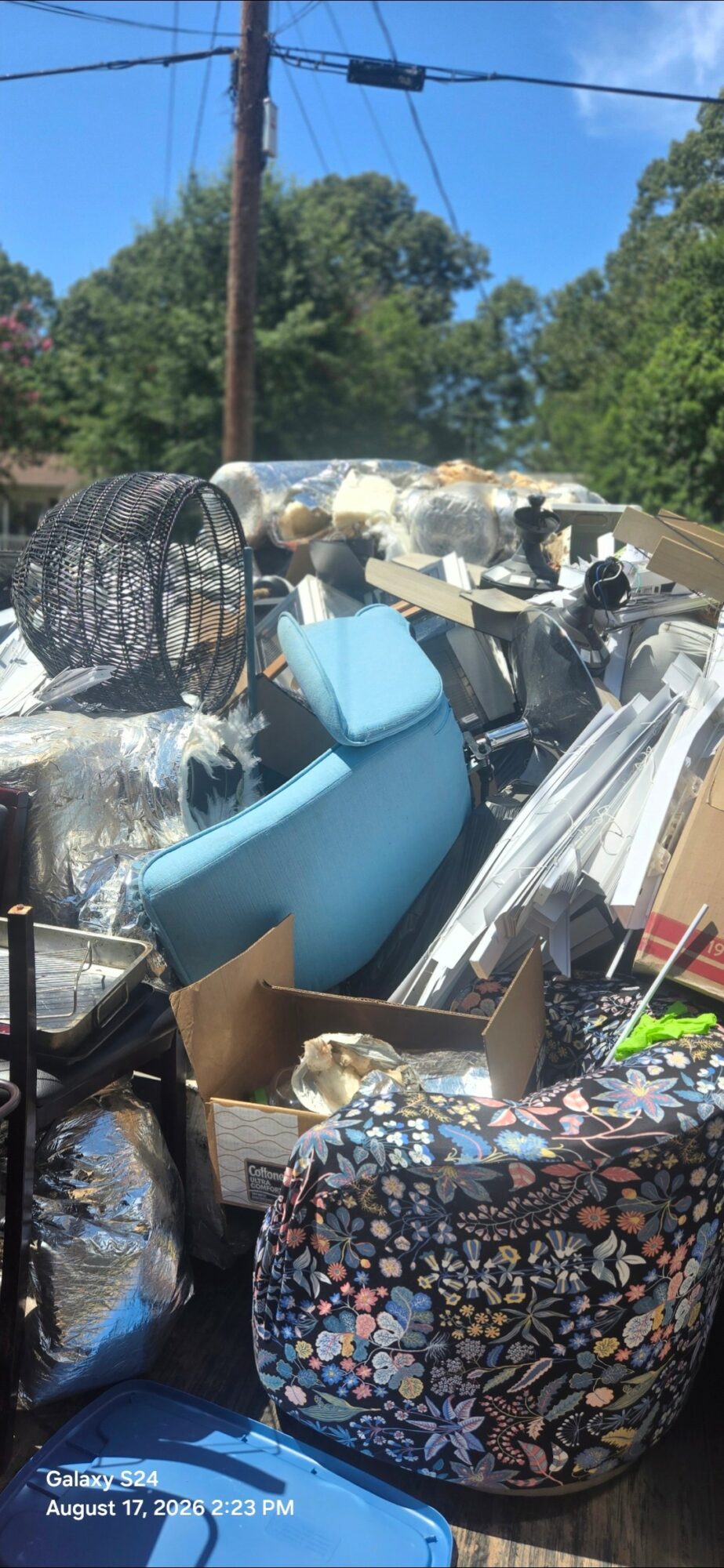 Pile of various items including bags, boxes, and furniture outdoors with trees and a utility pole in background.