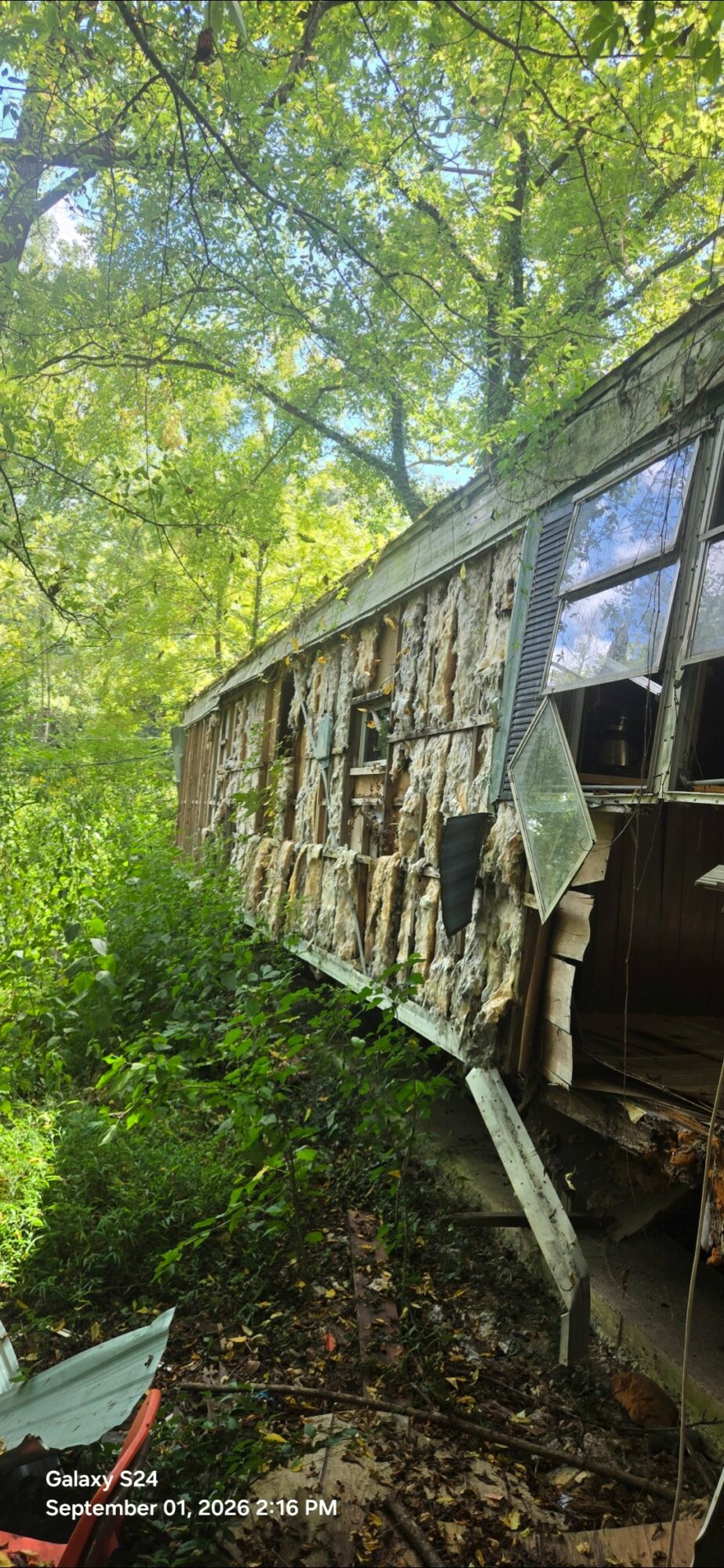 Old, damaged trailer with broken windows and peeling paint in a wooded area, surrounded by green foliage.