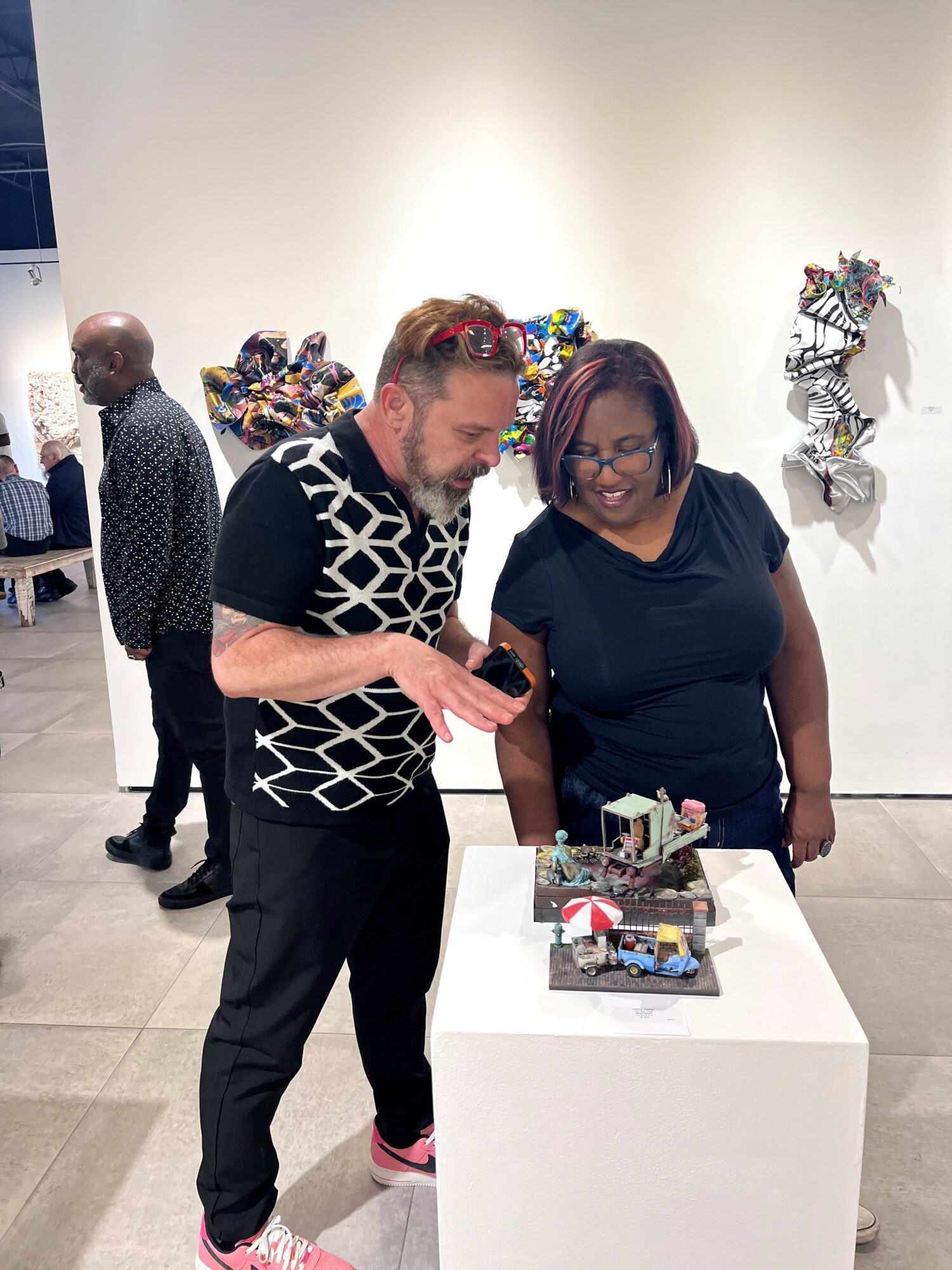 Two people look at a decorated cake on a white pedestal in an art gallery, with artwork on the wall behind them.