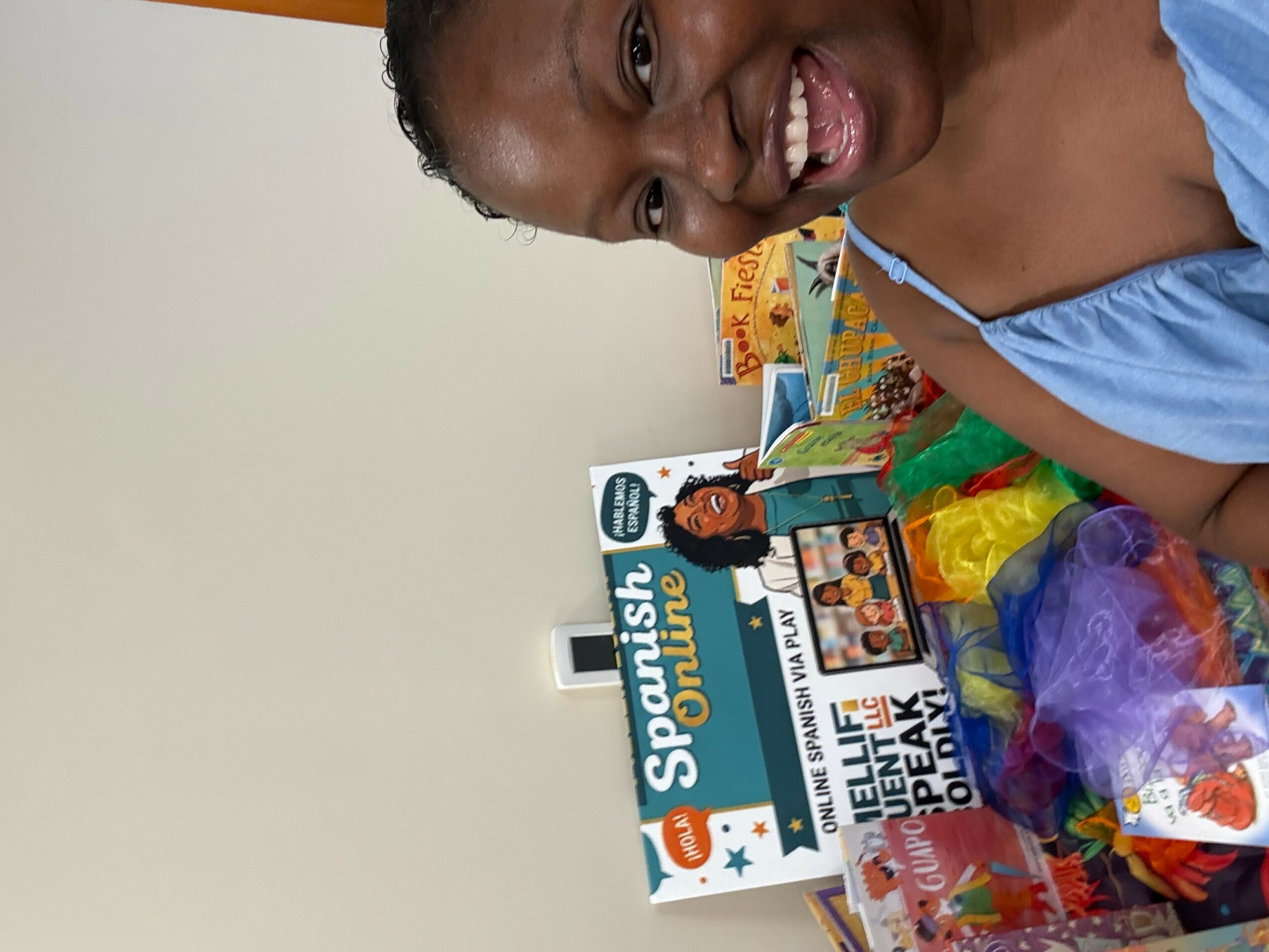 Smiling girl with dark hair and blue top, surrounded by colorful toys and items, against a plain background.
