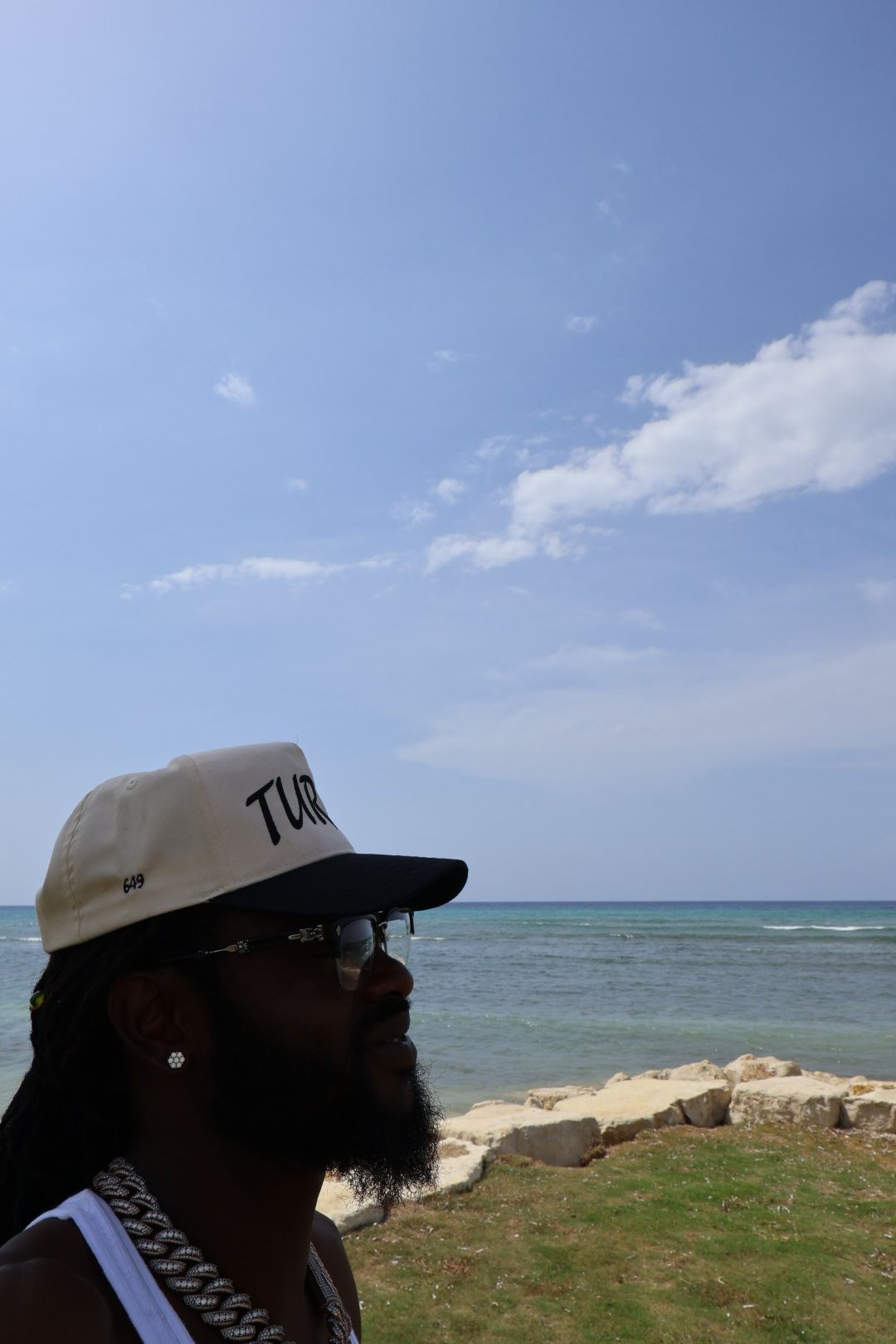 Person wearing a cap and sunglasses near water with a grassy area and rocks, sky with clouds in background.