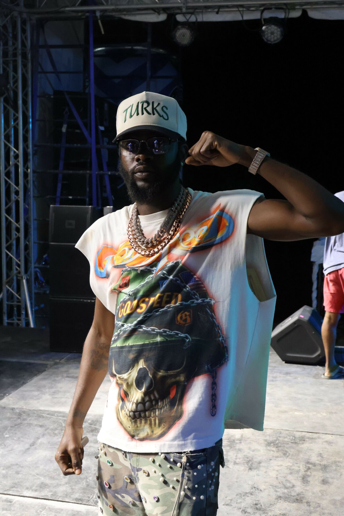 Person wearing a cap, graphic t-shirt, and camouflage pants, standing outdoors with a dark background.