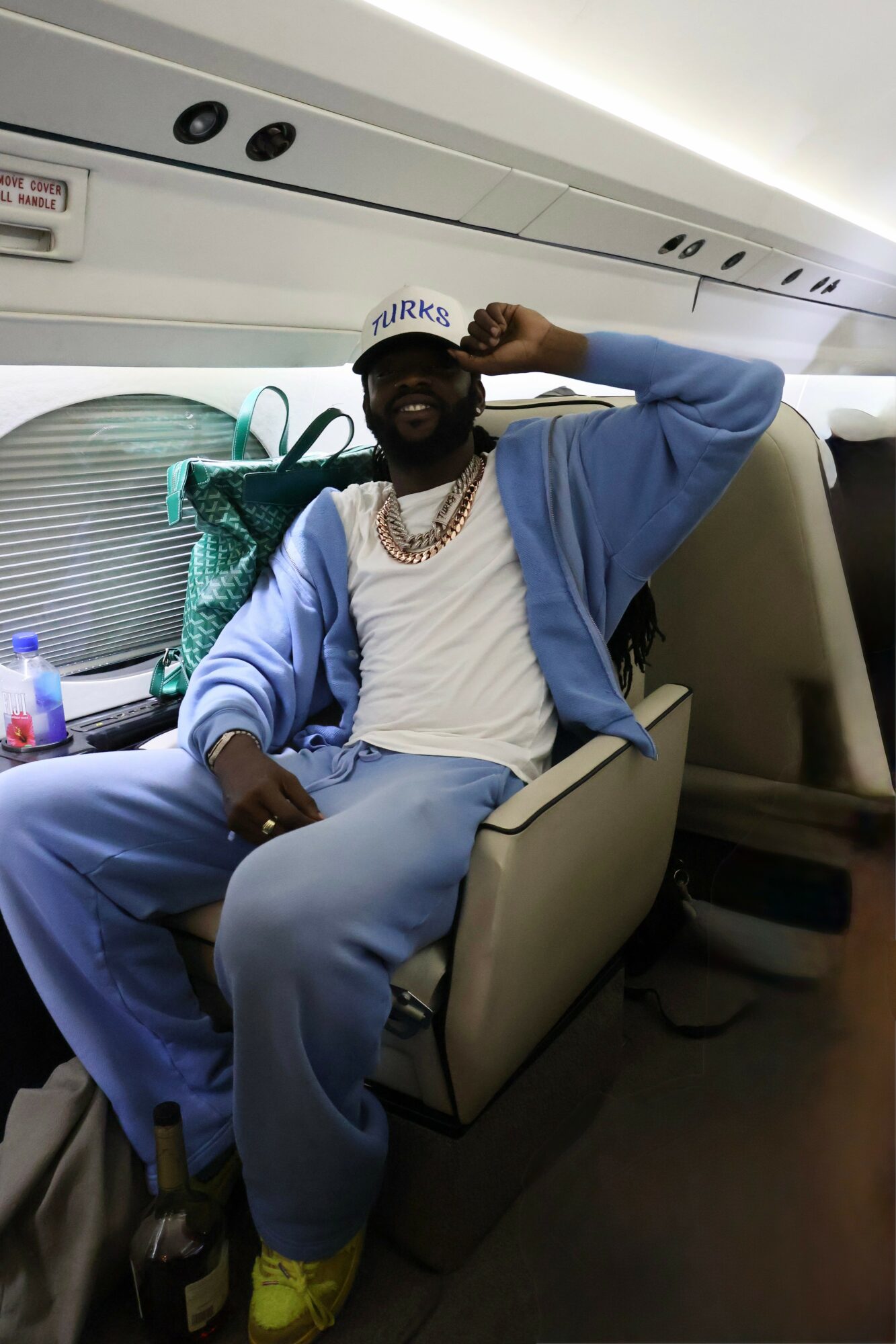 Person sitting on airplane seat, wearing a blue hoodie, white shirt, yellow shoes, and sunglasses, smiling and adjusting cap.