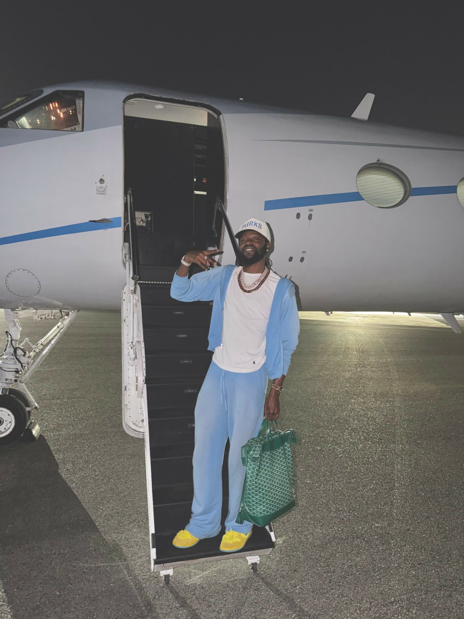 Person standing on stairs next to private jet at night, holding a green bag, wearing blue and white clothing, smiling.