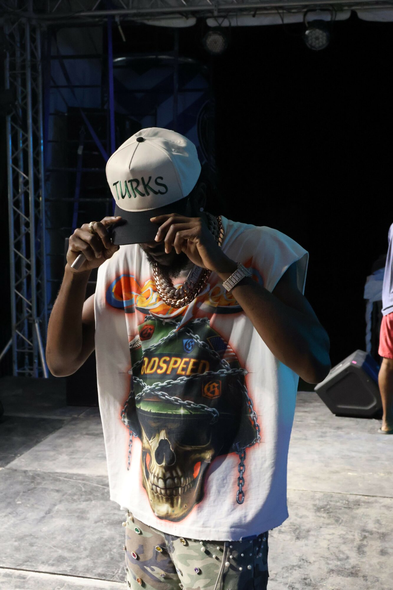 Person wearing a white helmet and a colorful T-shirt, holding a microphone, standing outdoors on a concrete surface.