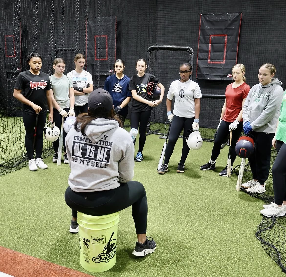 Group of young women listening to a coach in an indoor sports facility with green turf and equipment.