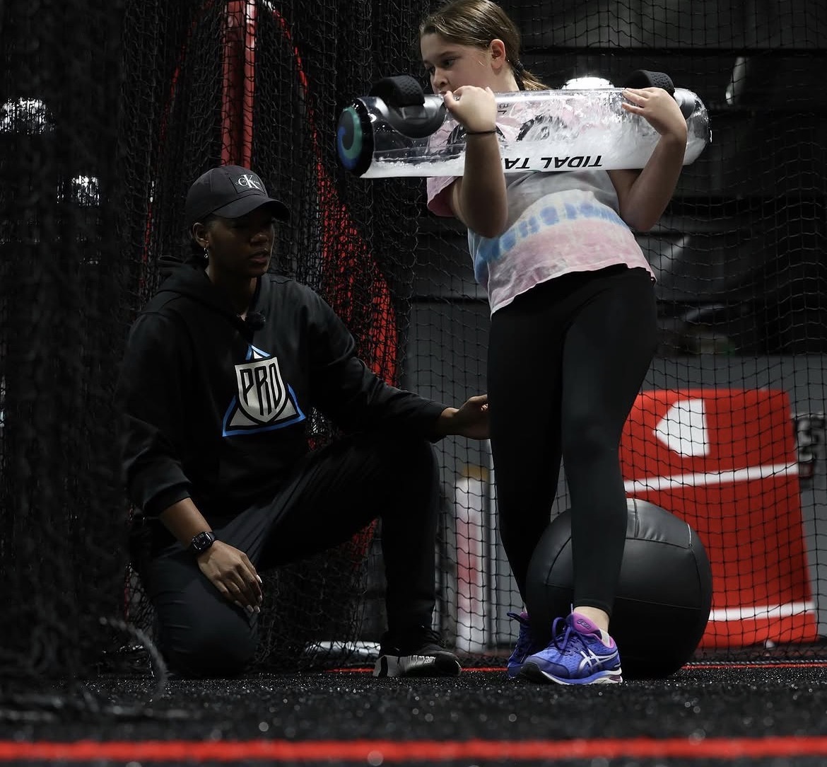 Young girl holding a baseball bat, with a coach kneeling beside her in a sports training facility.