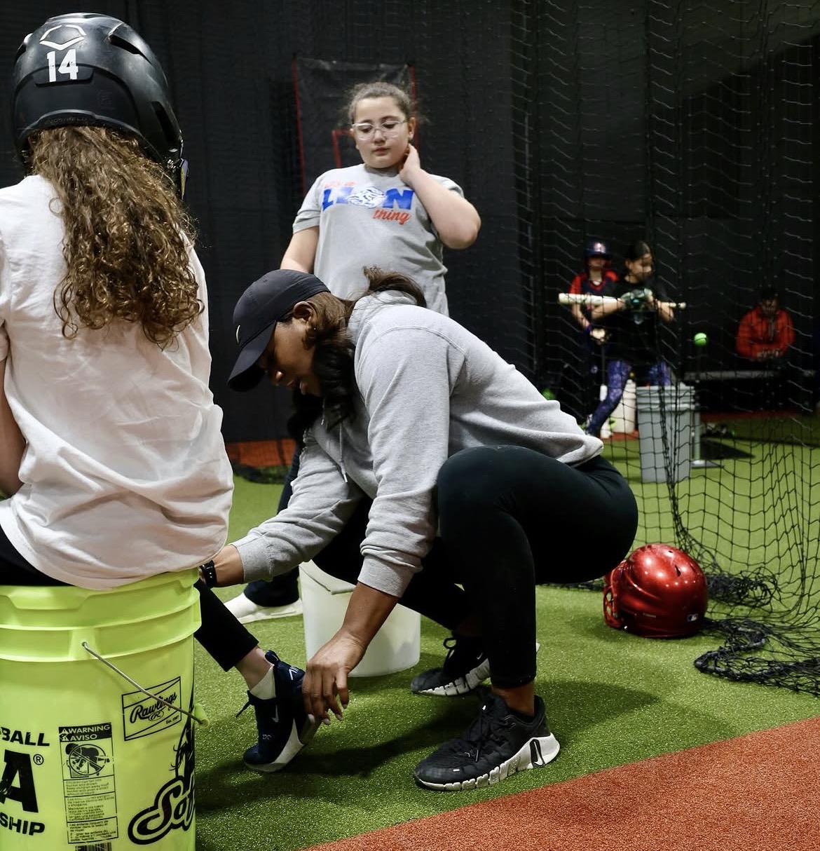 A woman helps a child put on a baseball glove in an indoor batting cage, with another girl watching in the background.