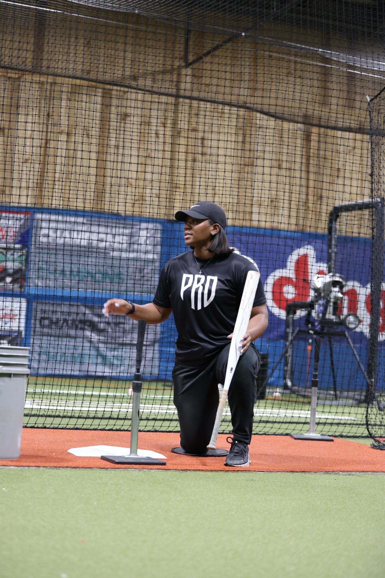 Person in black shirt and cap practicing baseball on a field with a fence background.