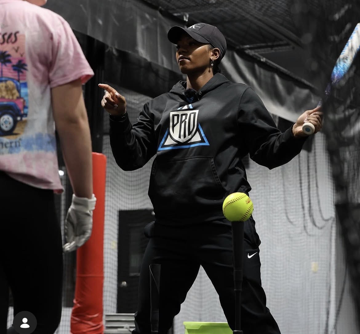 Woman in black hoodie and cap holding a tennis ball, with a person in a pink shirt nearby, indoor sports setting.