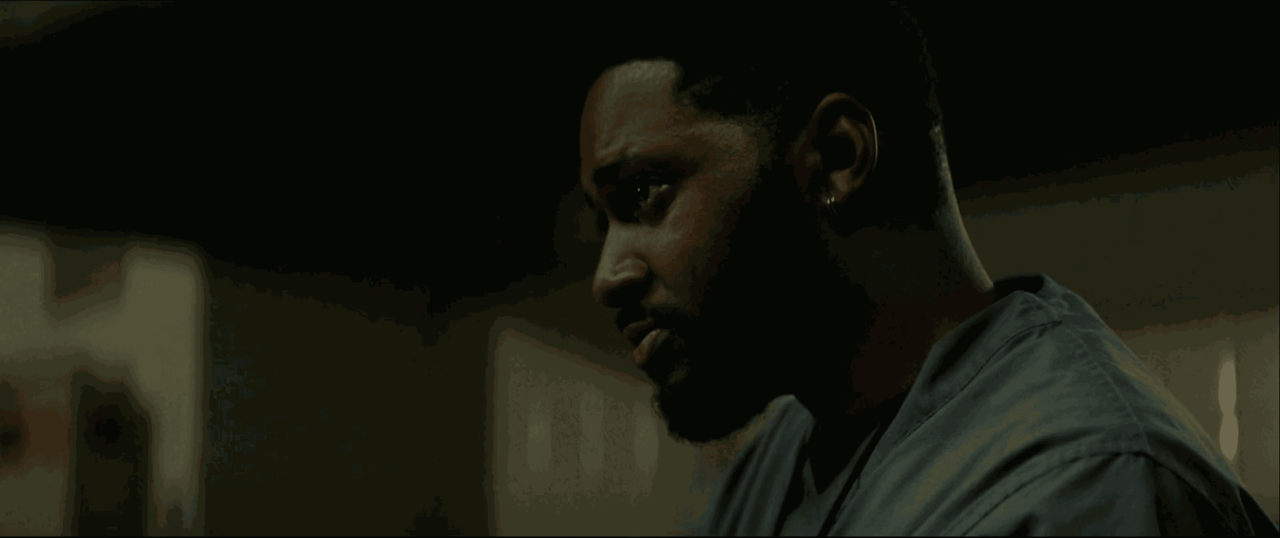 Side profile of a man with dark hair and beard, looking downward, in a dimly lit room.