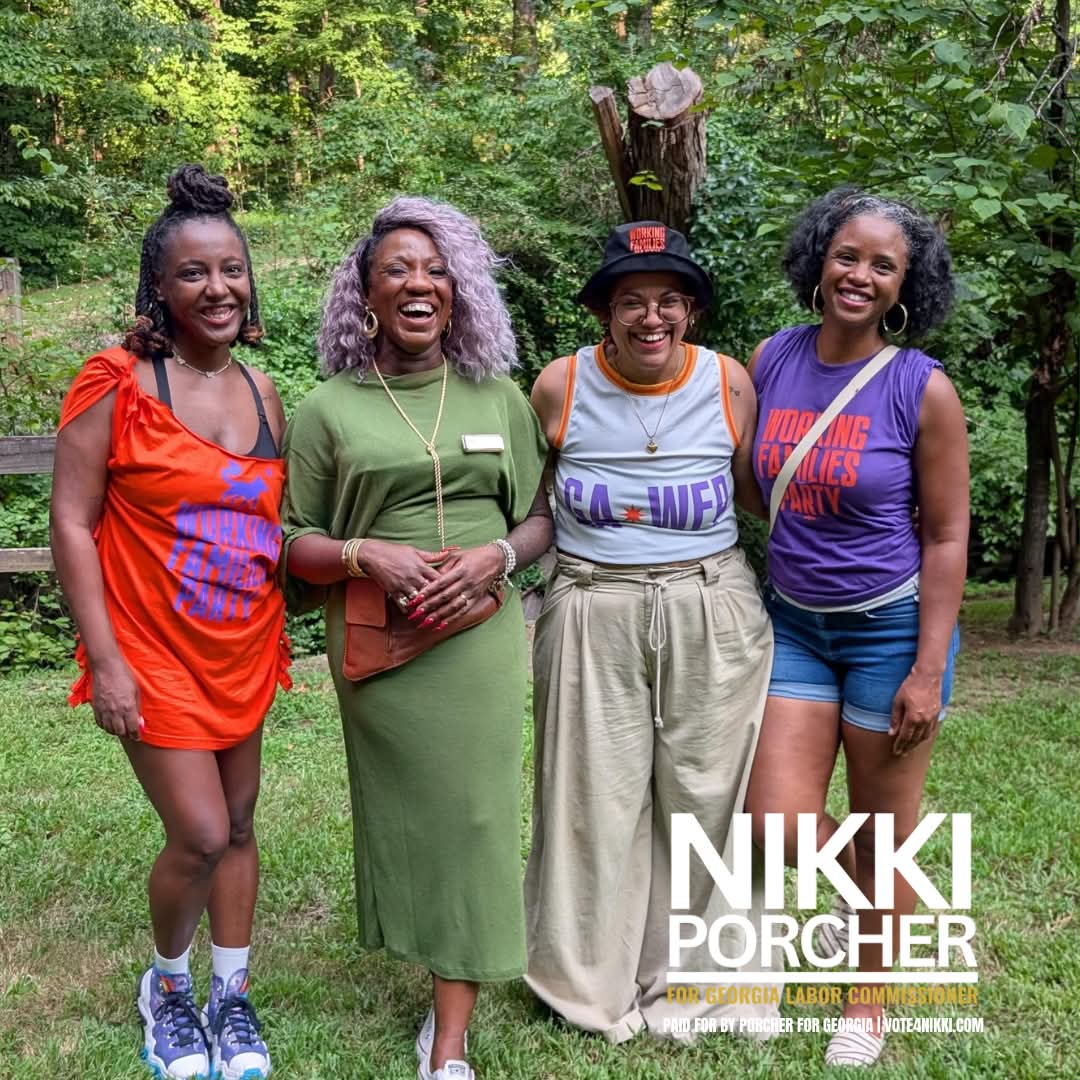 Four women standing outdoors in a wooded area, smiling, wearing colorful casual clothing, with trees in the background.