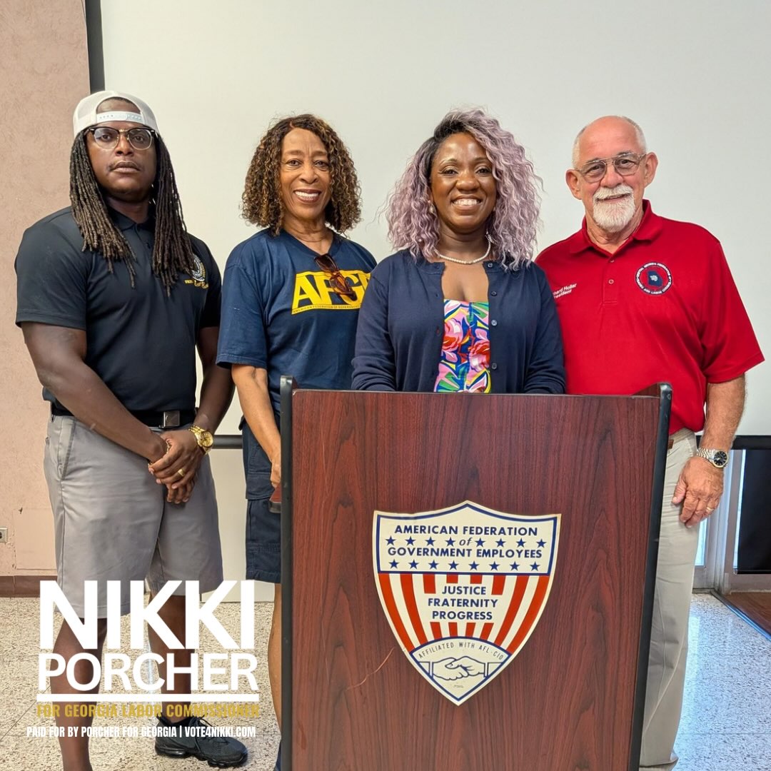 Four people standing behind a podium with a shield emblem, smiling, in a room with a white wall.