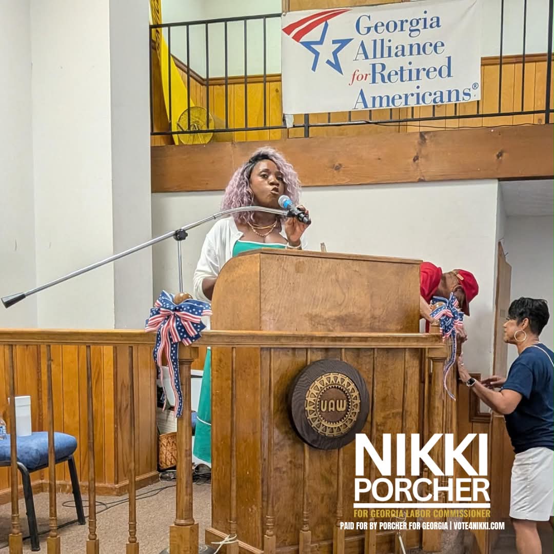 Woman speaking at a podium with a Georgia Alliance banner above, decorated with ribbons, in a room with wooden paneling.