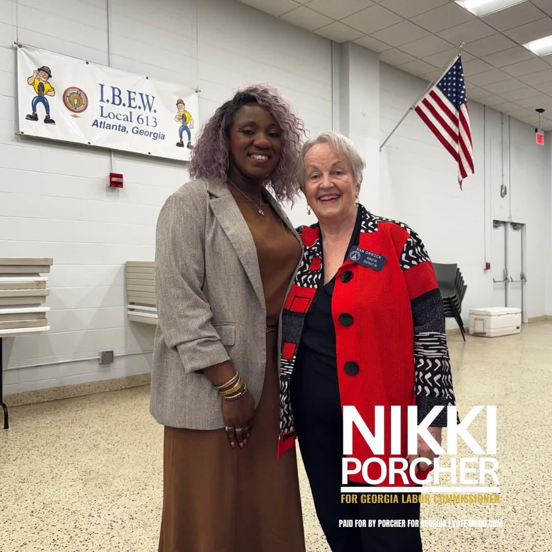 Two women smiling indoors, one wearing a beige blazer and the other a red and black patterned jacket, with an American flag and a banner in the background.