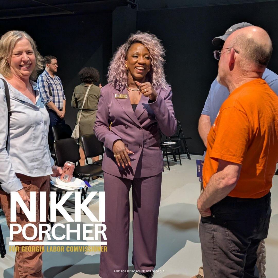 Three people smiling and talking in a room with chairs and other attendees in the background.