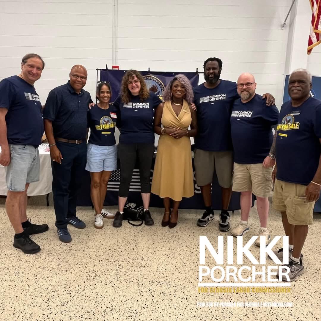 Group of eight people standing indoors, some wearing matching navy shirts, smiling, with a banner behind them, and a white wall.
