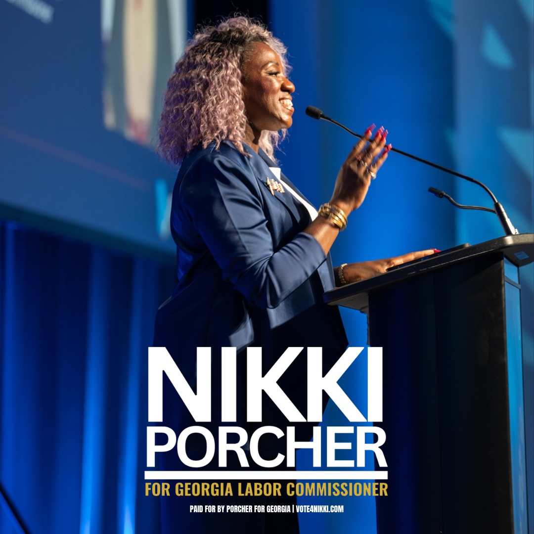 Woman speaking at a podium with a blue background, wearing a dark blazer and gold jewelry, smiling and gesturing with her hands.