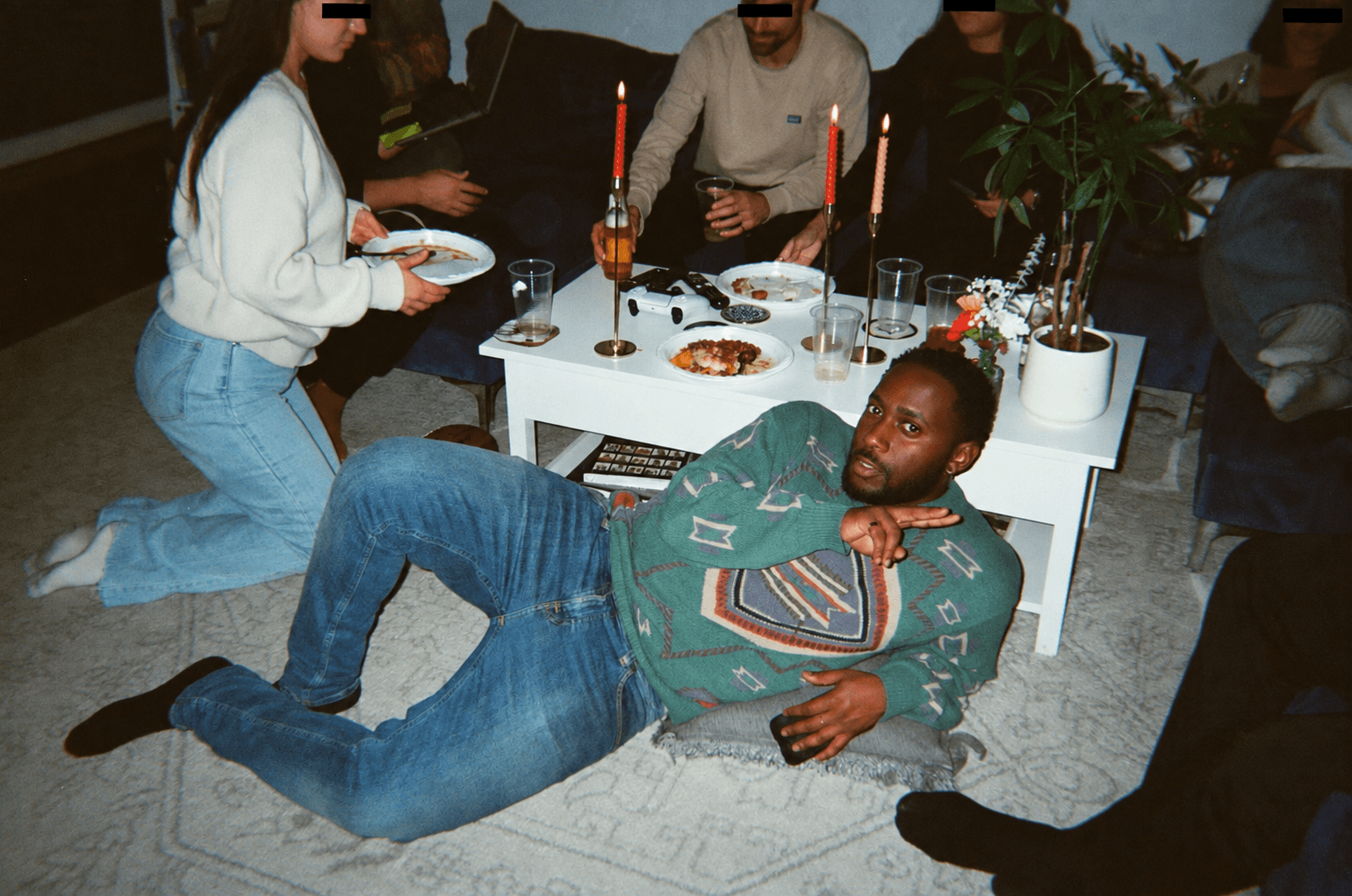 Man lying on the floor in front of a table with drinks and snacks, with people standing and sitting around.