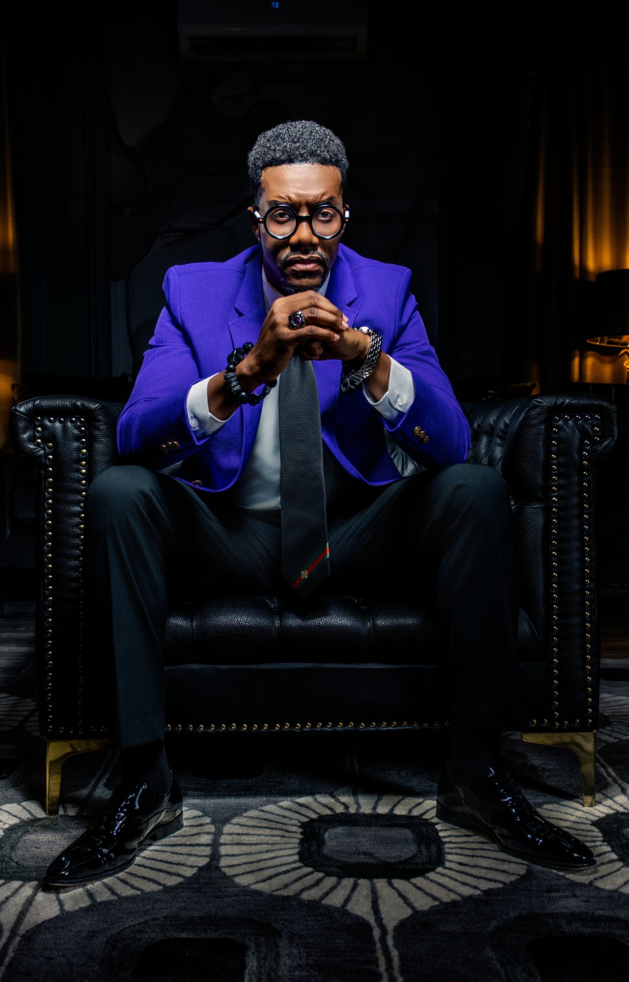 Man in blue blazer and glasses sitting on a black leather armchair with hands clasped, dark background, dim lighting.