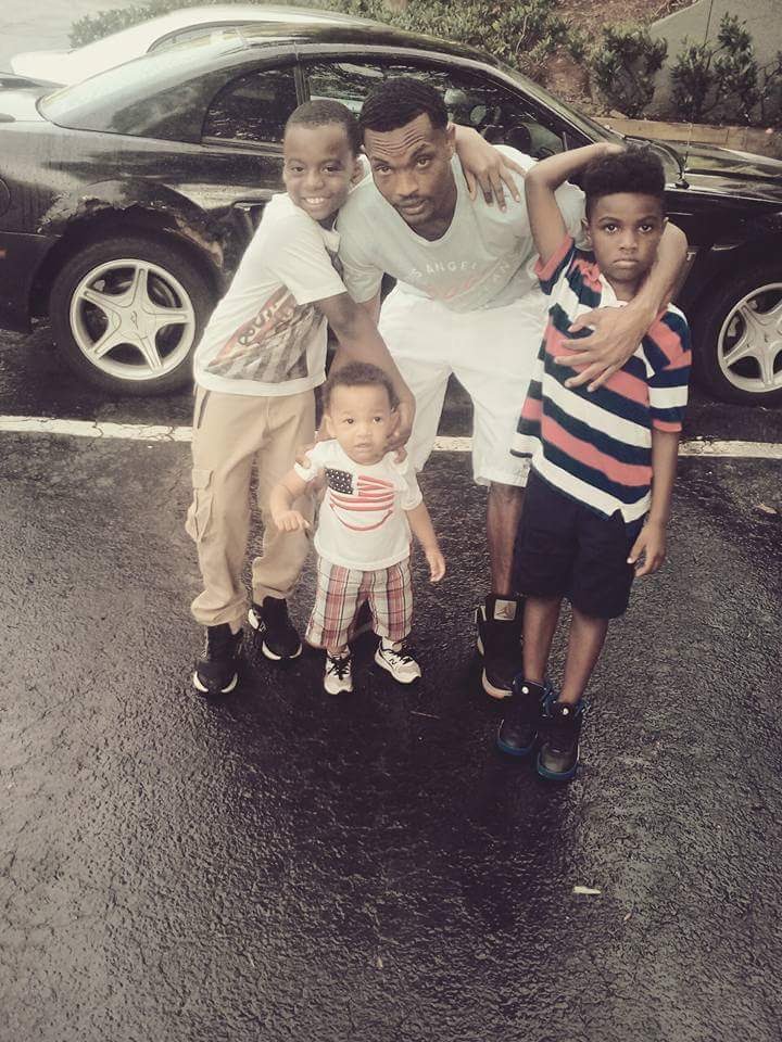 Four children and a man pose in a parking lot with a black car behind them, smiling and hugging each other.