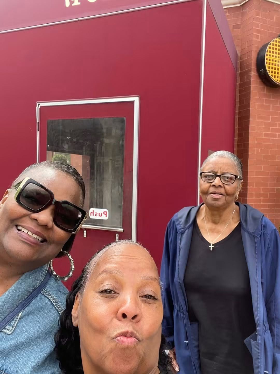Three women standing outside a red building, smiling and posing for a photo.