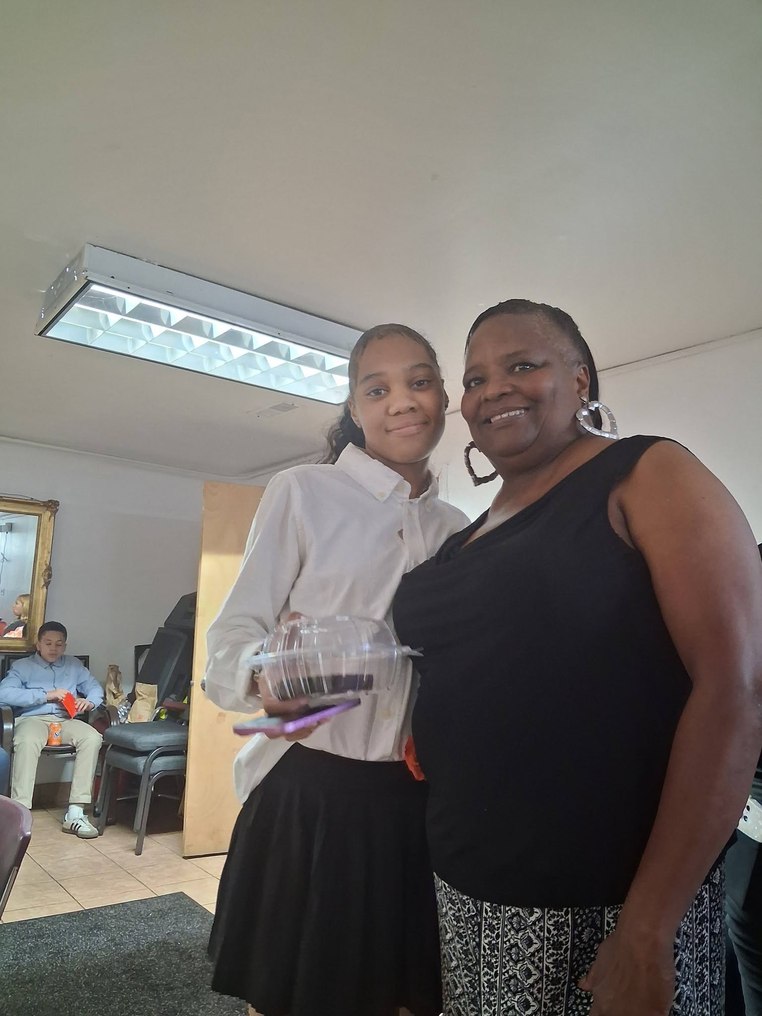Two women standing indoors, one holding a clear container, smiling at the camera.