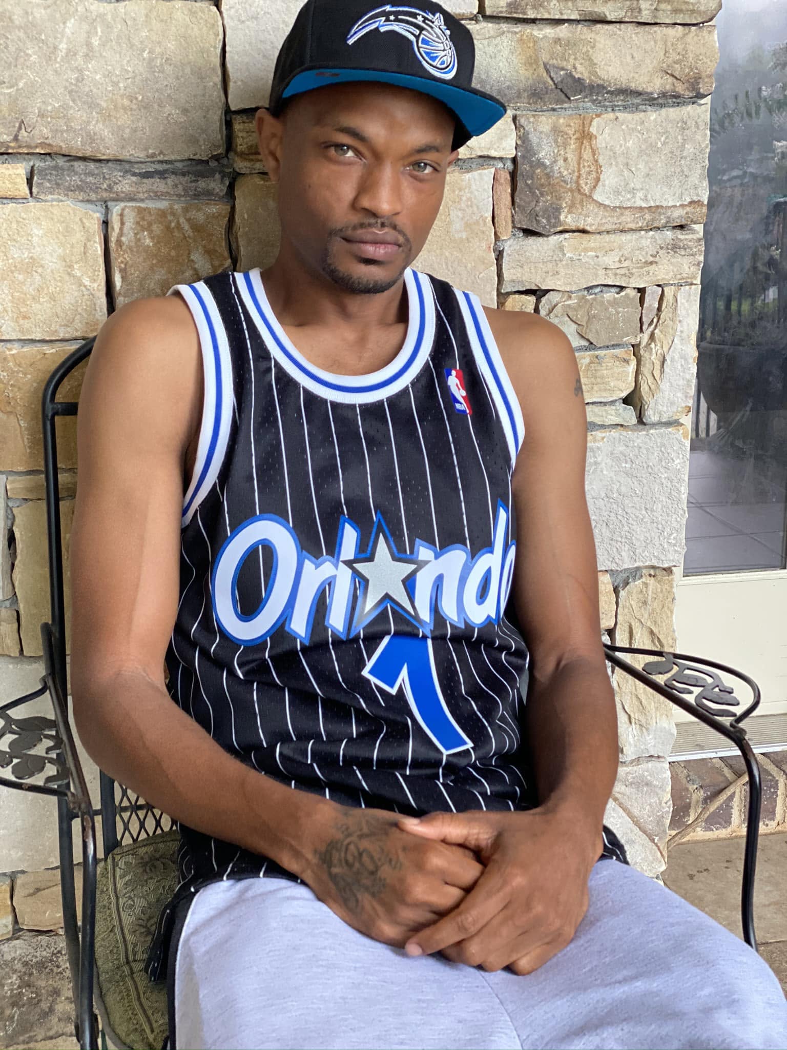Man sitting outdoors against a stone wall, wearing a black and blue basketball jersey and a cap.