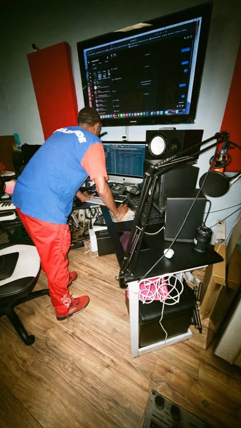 Person in blue shirt and red pants working with audio equipment in a room with a large monitor, speakers, and cables.